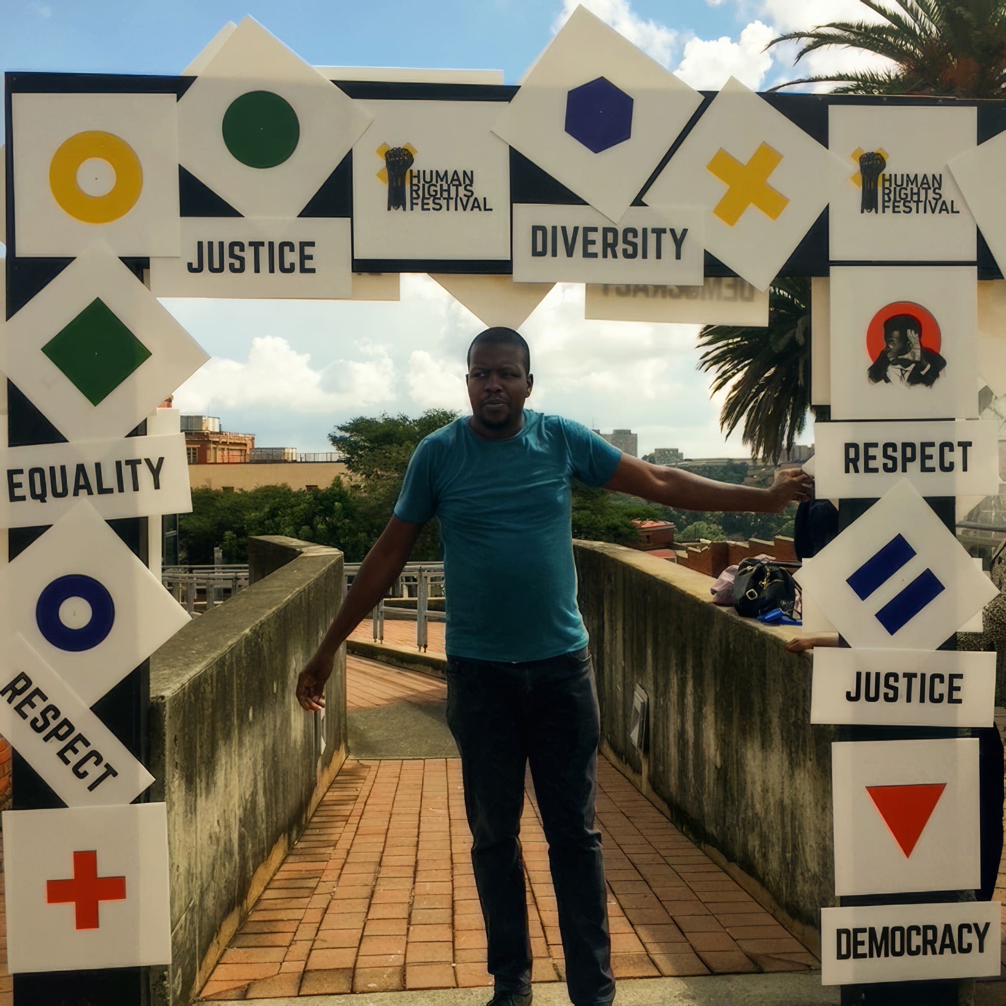 A man in a teal shirt stands under a festival archway displaying square signs with words like “Diversity,” “Justice,” “Equality,” “Respect,” and “Democracy,” along with geometric symbols. The scene is outdoors under a partly cloudy sky.