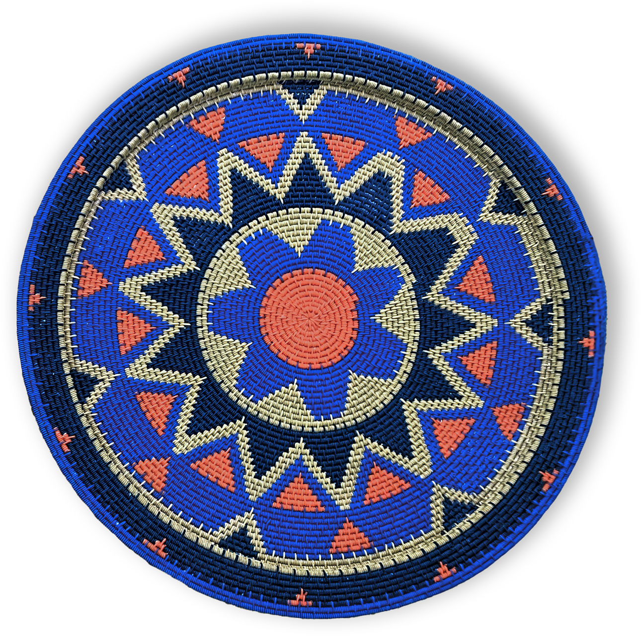 A round, woven basket lid features a geometric pattern in blue, coral orange, cream, and navy. The design includes a central flower-like shape, surrounded by alternating triangles and zigzag bands, creating a vibrant, symmetrical motif.