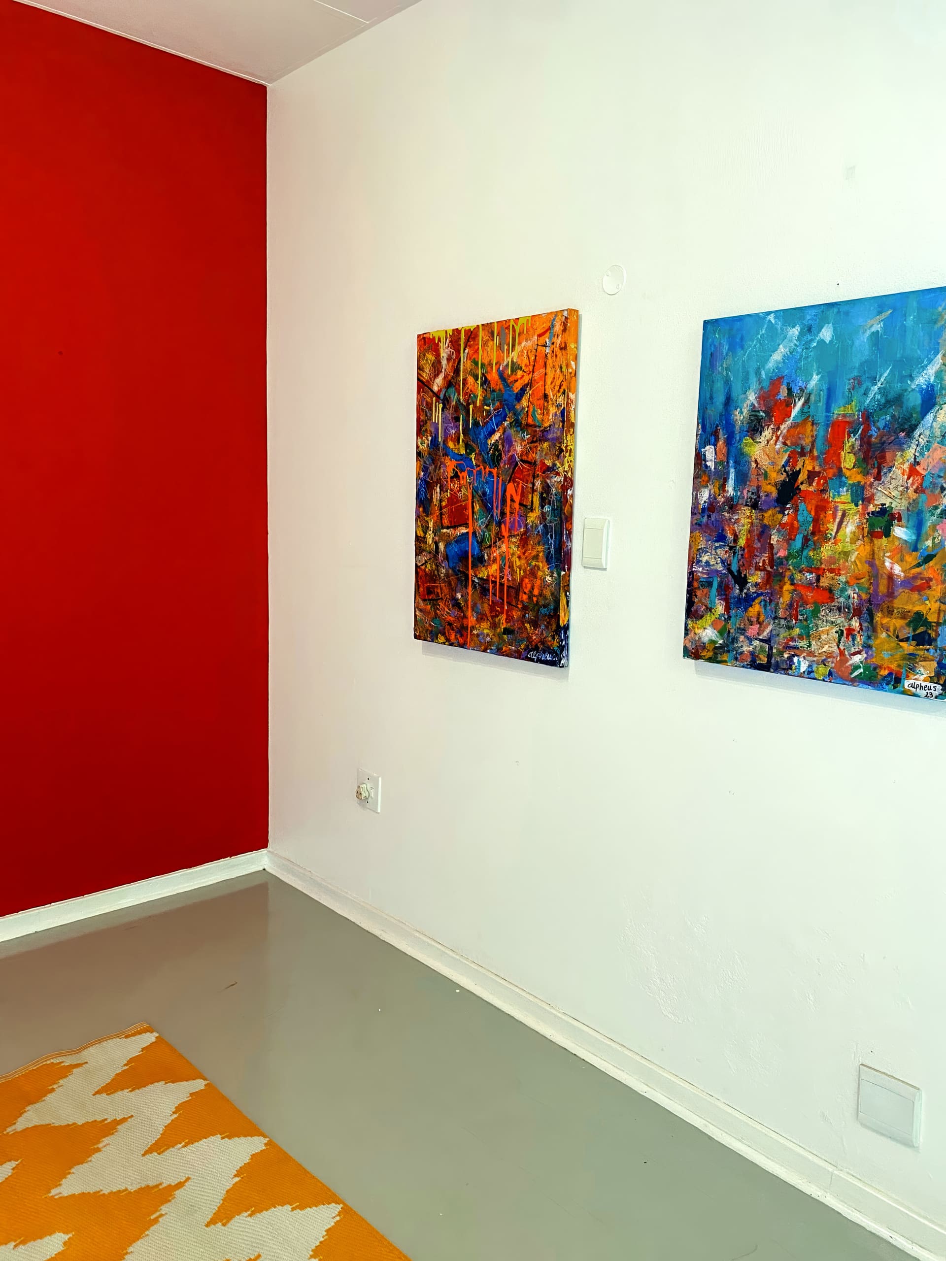 A modern room with a red feature wall and white adjoining wall. Two colourful abstract paintings hang side by side on the white wall. The floor is grey, and a yellow and white chevron rug covers part of it. The room appears minimal and artistic.