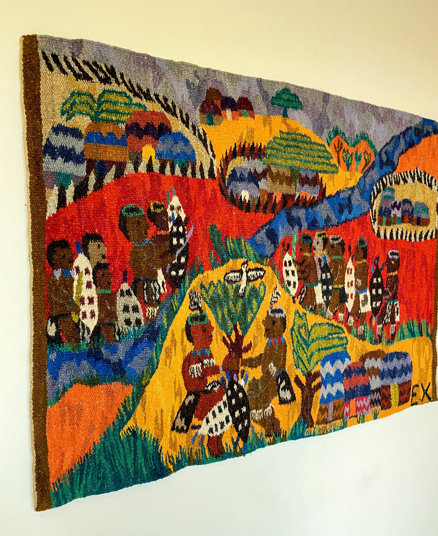 A colourful woven tapestry hangs on a wall, depicting groups of people in patterned attire, huts, trees, and hills in bold reds, yellows, blues, and greens. The scene is lively and stylised, showcasing traditional village life and culture.
