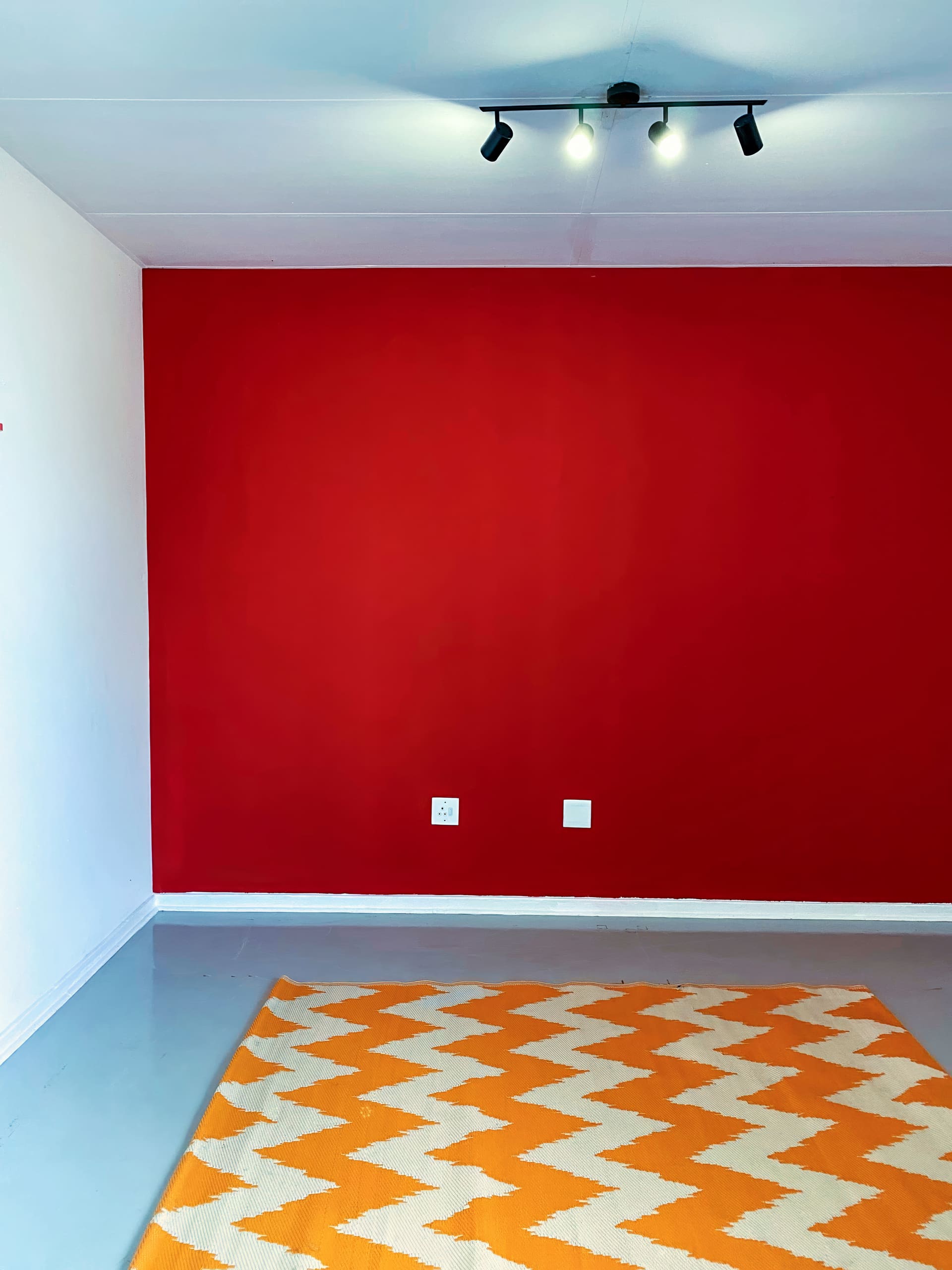 A minimalist room with a bold red feature wall, white side walls, grey floor, and a geometric orange-and-white rug. Two electrical sockets are visible on the red wall beneath a black four-bulb ceiling light fitting. The room is empty.