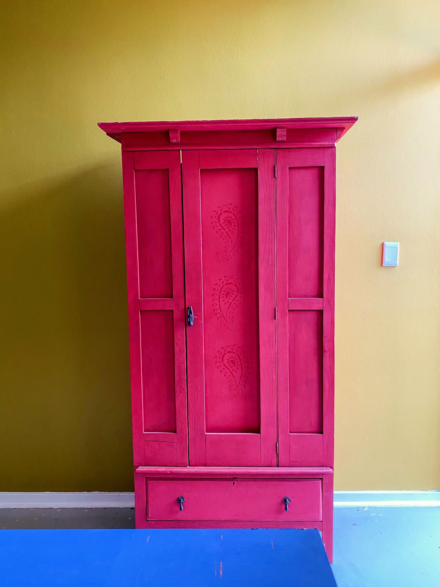 A tall, vibrant pink wooden cupboard with a single door and a large bottom drawer stands against a mustard yellow wall. The door features faint paisley patterns. The floor is blue, and there is a small light switch on the wall to the right.