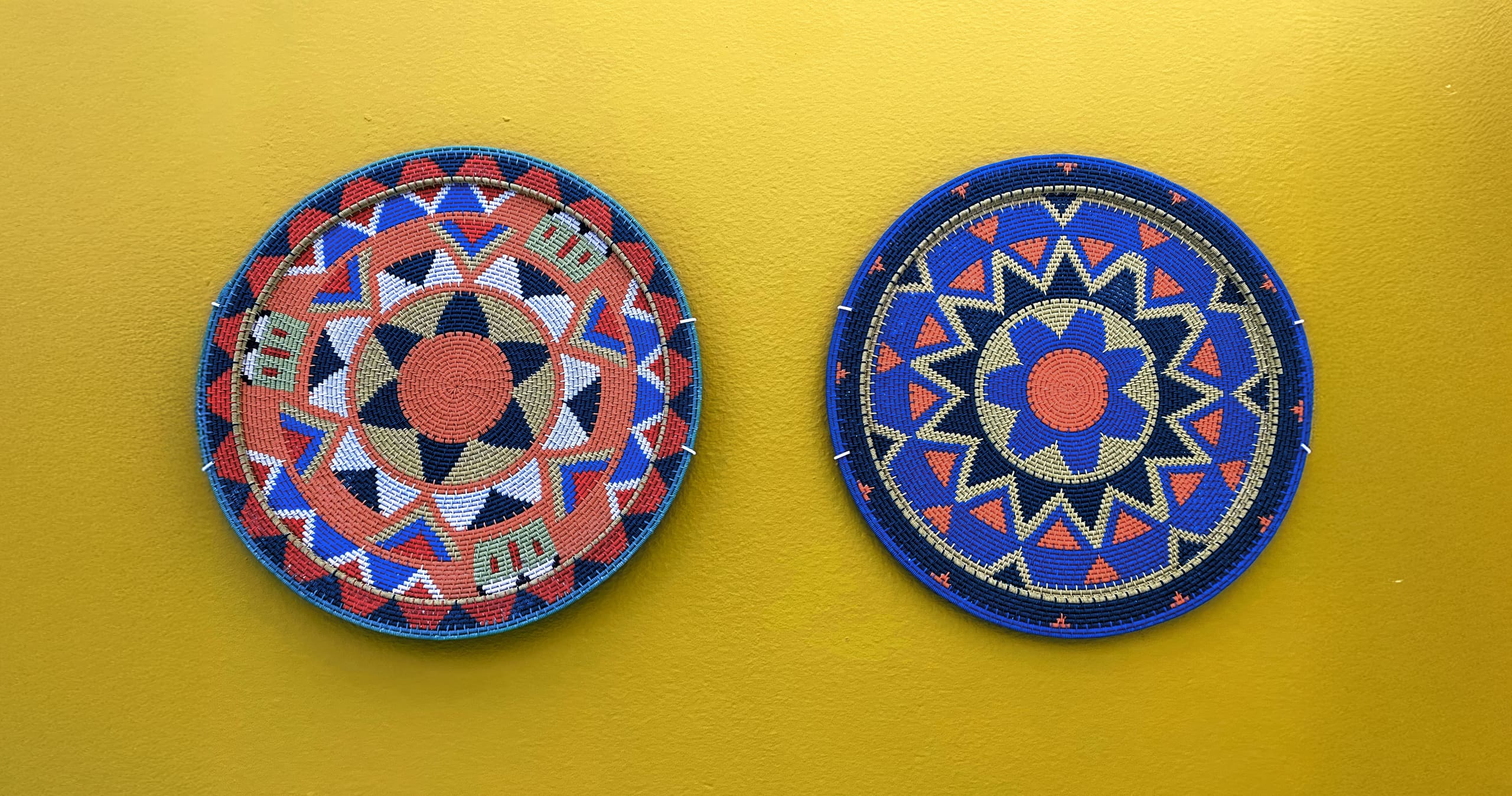 Two round, woven baskets with intricate geometric patterns hang side by side on a yellow wall. The left basket features red, blue, black, and white designs, whilst the right basket displays blue, red, yellow, and white star-like shapes.