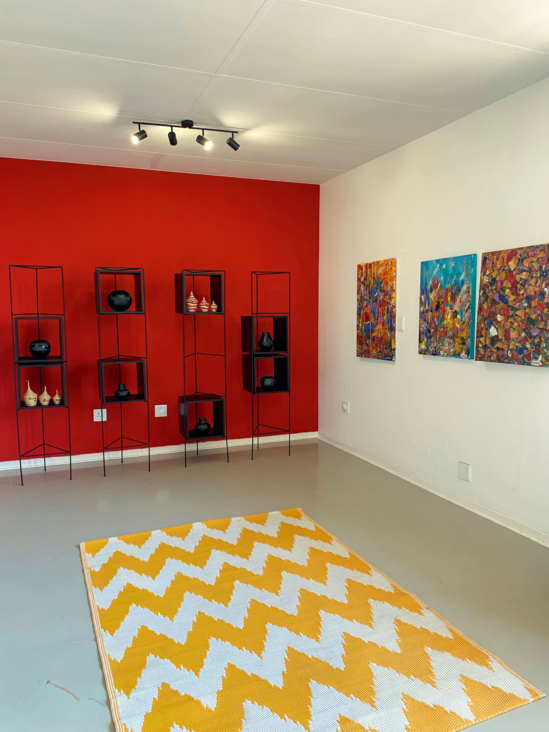 A modern art gallery with a red feature wall featuring black metal shelves displaying ceramic vases. Three colourful abstract paintings hang on a white wall. The floor has a yellow and white zigzag patterned rug, and track lighting is installed on the ceiling.