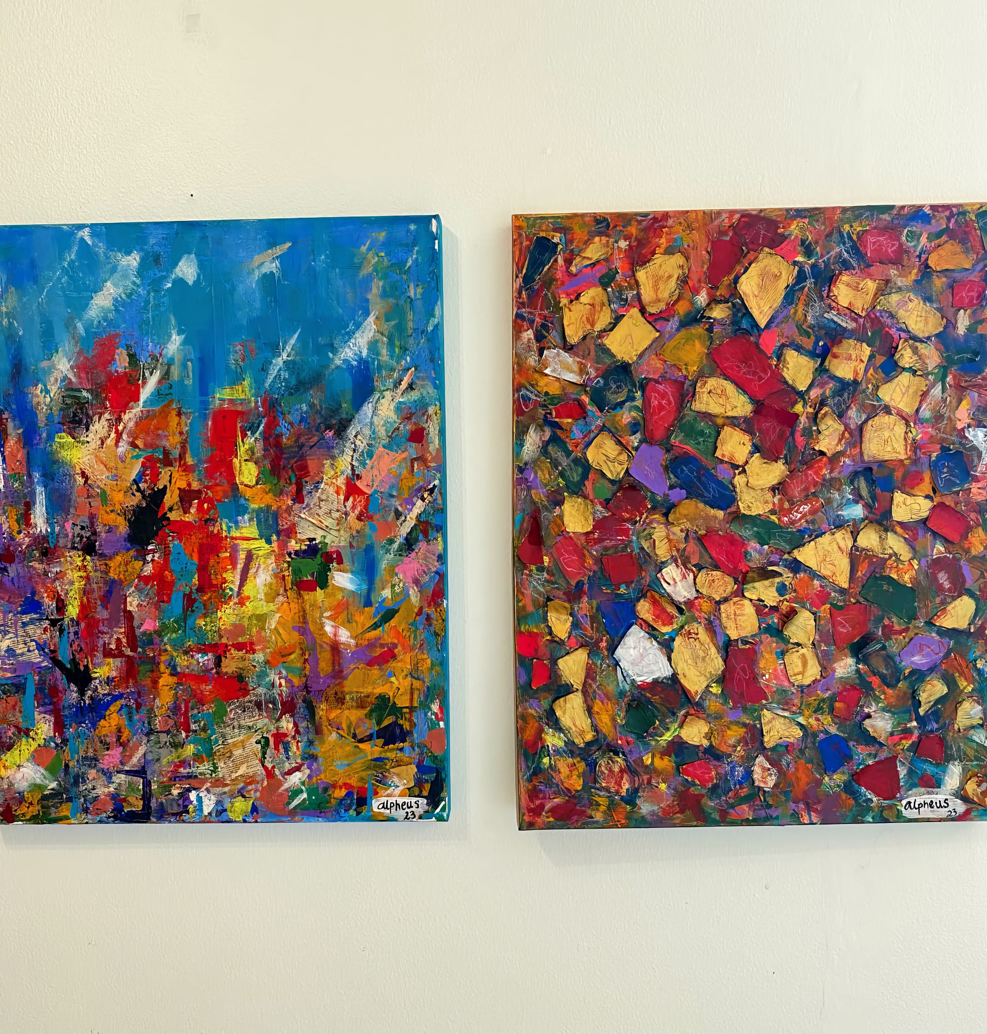 Two abstract paintings hang side by side on a white wall. The left artwork features vibrant blue, red, orange, and yellow brushstrokes, whilst the right piece displays a dense, mosaic pattern of red, yellow, green, and gold with bold, irregular shapes.