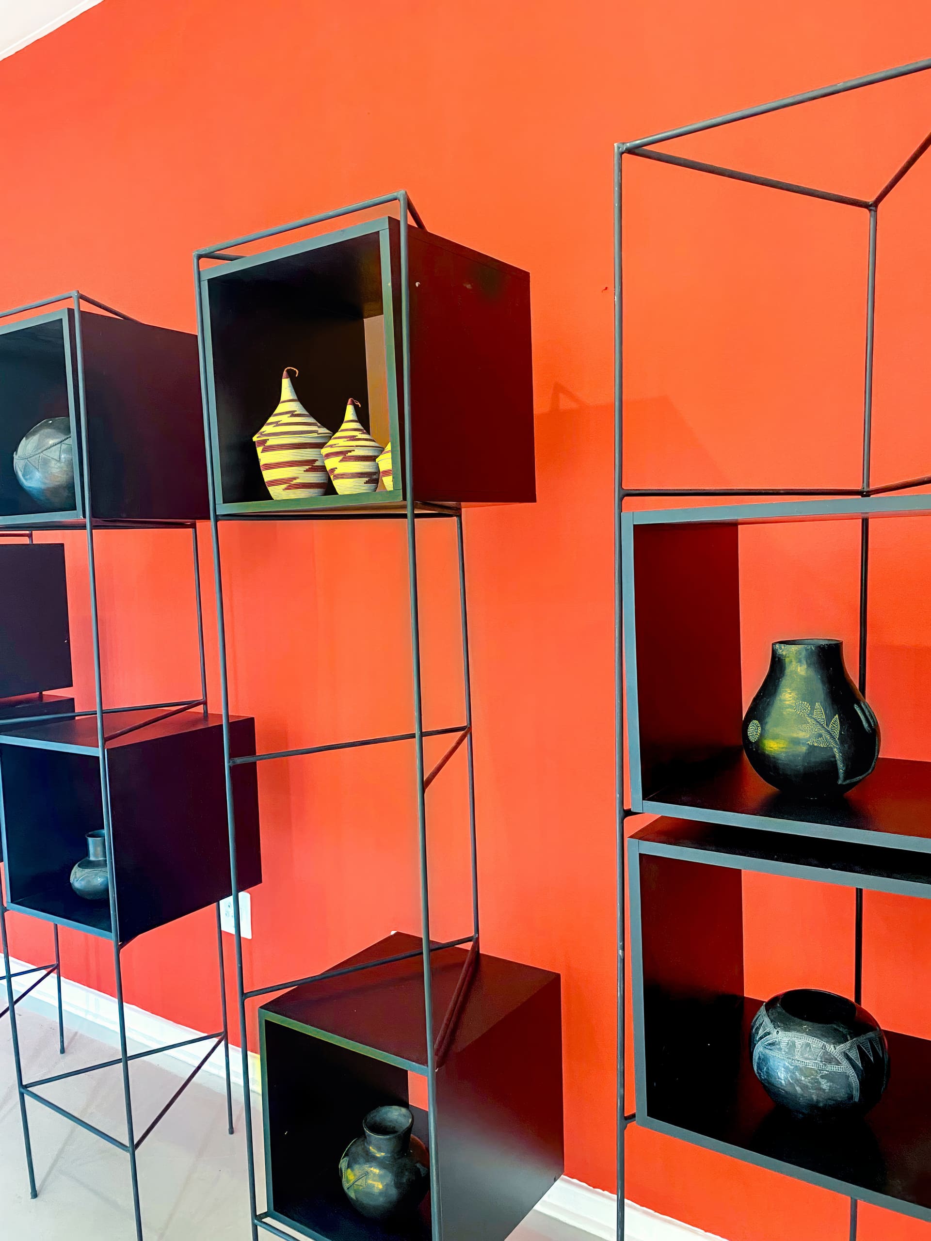 Black metal and wood display shelves stand against a bold red wall. The shelves hold various pottery pieces, including a pair of yellow striped vessels, a round dark vase, and other uniquely shaped ceramic pots in earthy tones.