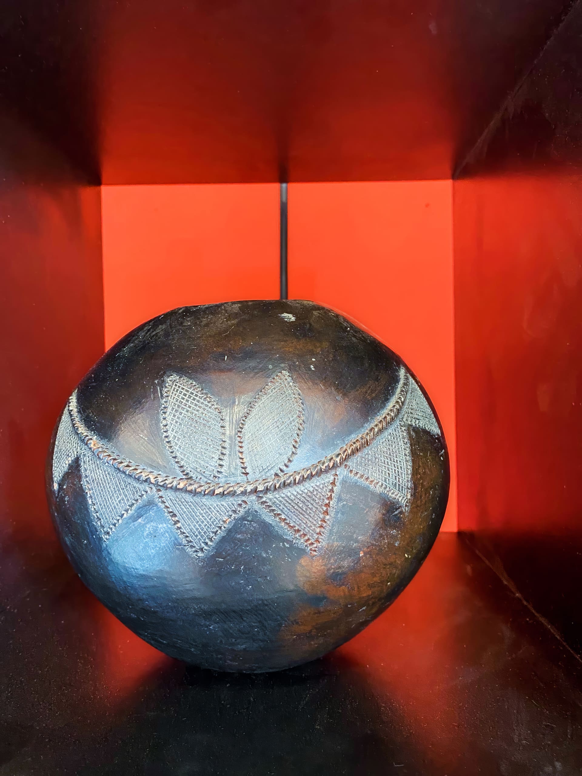 A round, dark brown pottery vessel with carved geometric and leaf-like patterns sits on a black shelf against a bold orange-red background. The pot's surface shows signs of wear and age, highlighting its hand-crafted texture and intricate decoration.