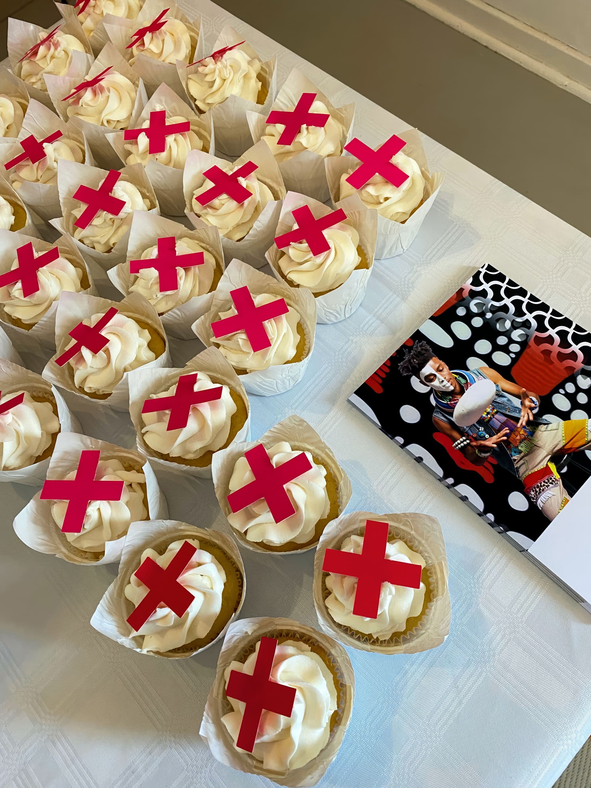 A group of cupcakes with swirled white icing, each topped with a red “X” decoration, sits on a white tablecloth next to a colourful magazine featuring a man in a playful costume and bold patterns.