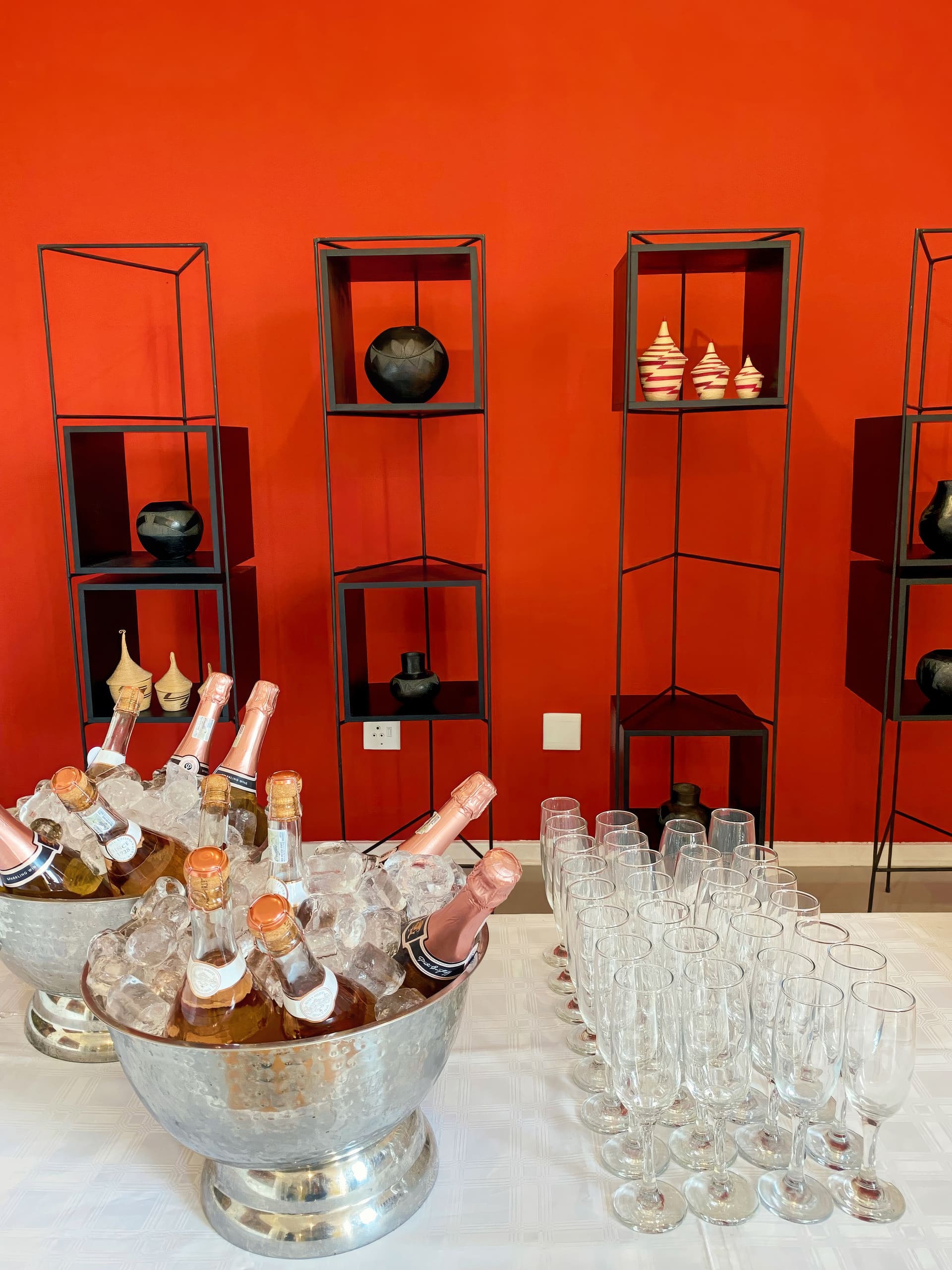 A table with bottles of rosé champagne in ice buckets and many empty champagne glasses in front of red shelves displaying decorative black and cream pottery pieces, set against a plain red wall.