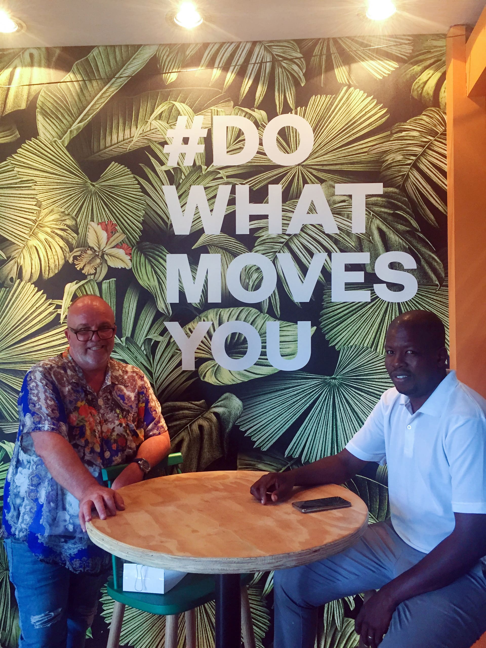 Two men sit at a round wooden table in front of a green leafy wall mural with large white text reading “#DO WHAT MOVES YOU.” One man wears a blue floral shirt and glasses; the other wears a white polo shirt. Both are smiling.