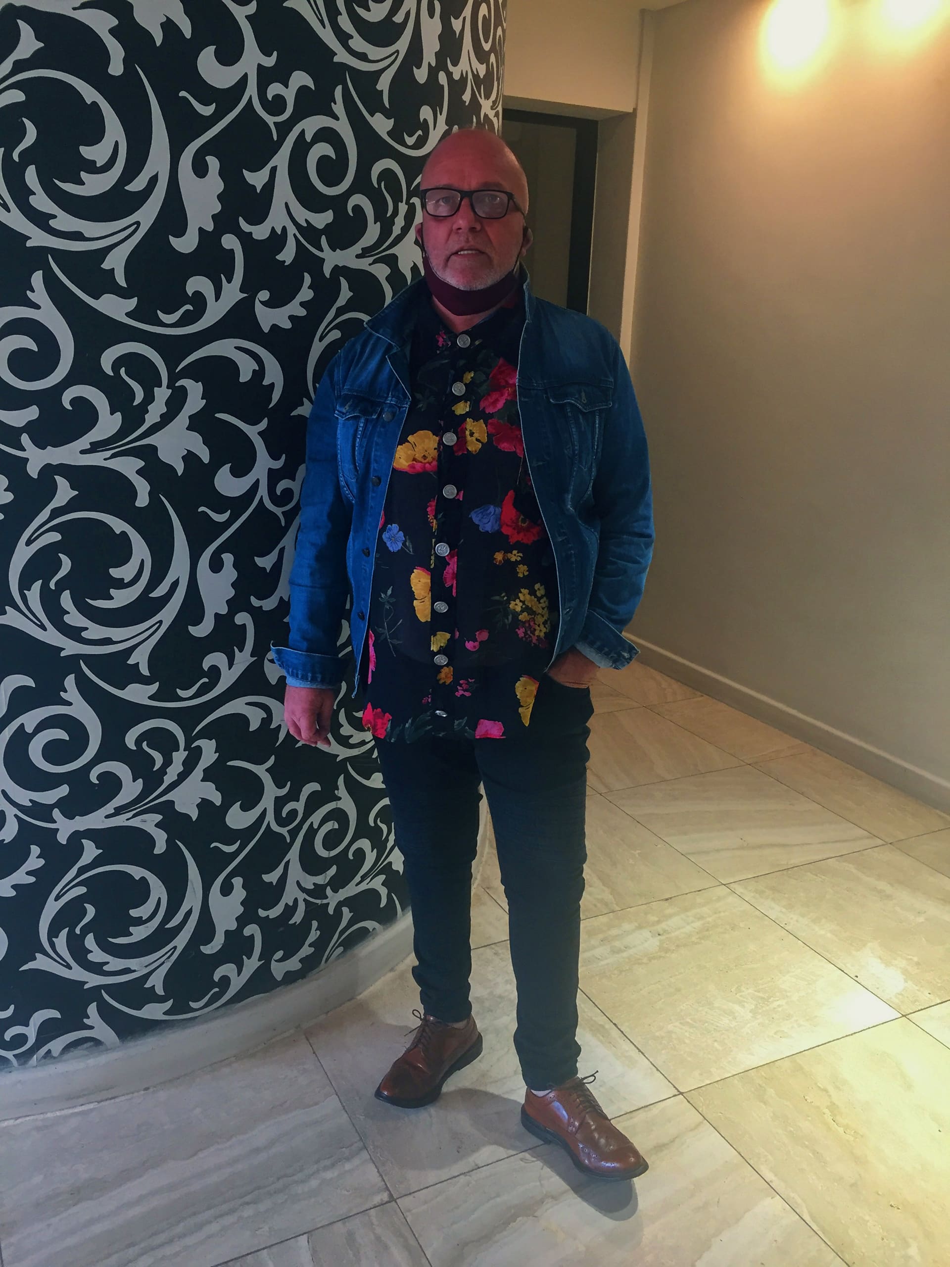 A man stands indoors on beige tiled flooring, next to a black and white ornate patterned wall. He wears glasses, a blue denim jacket, a black shirt with bright floral prints, dark trousers, and brown shoes. One hand is in his pocket and he looks at the camera.