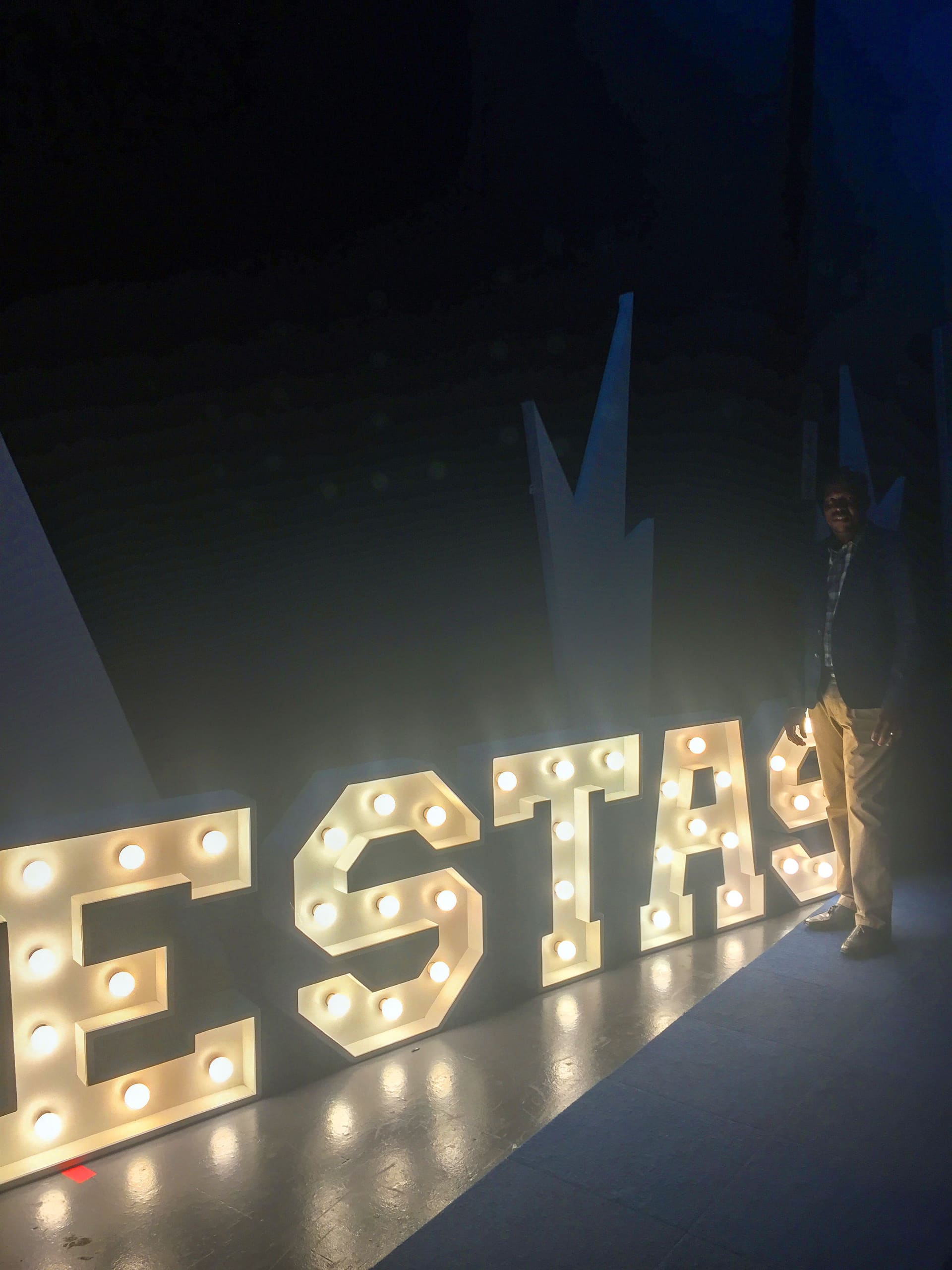 A person stands beside large, illuminated marquee letters spelling “ESTAS” on a dark floor. The area is dimly lit except for the warm glow of the letters. Abstract, angular decorations are visible in the background, adding to the dramatic lighting effect.