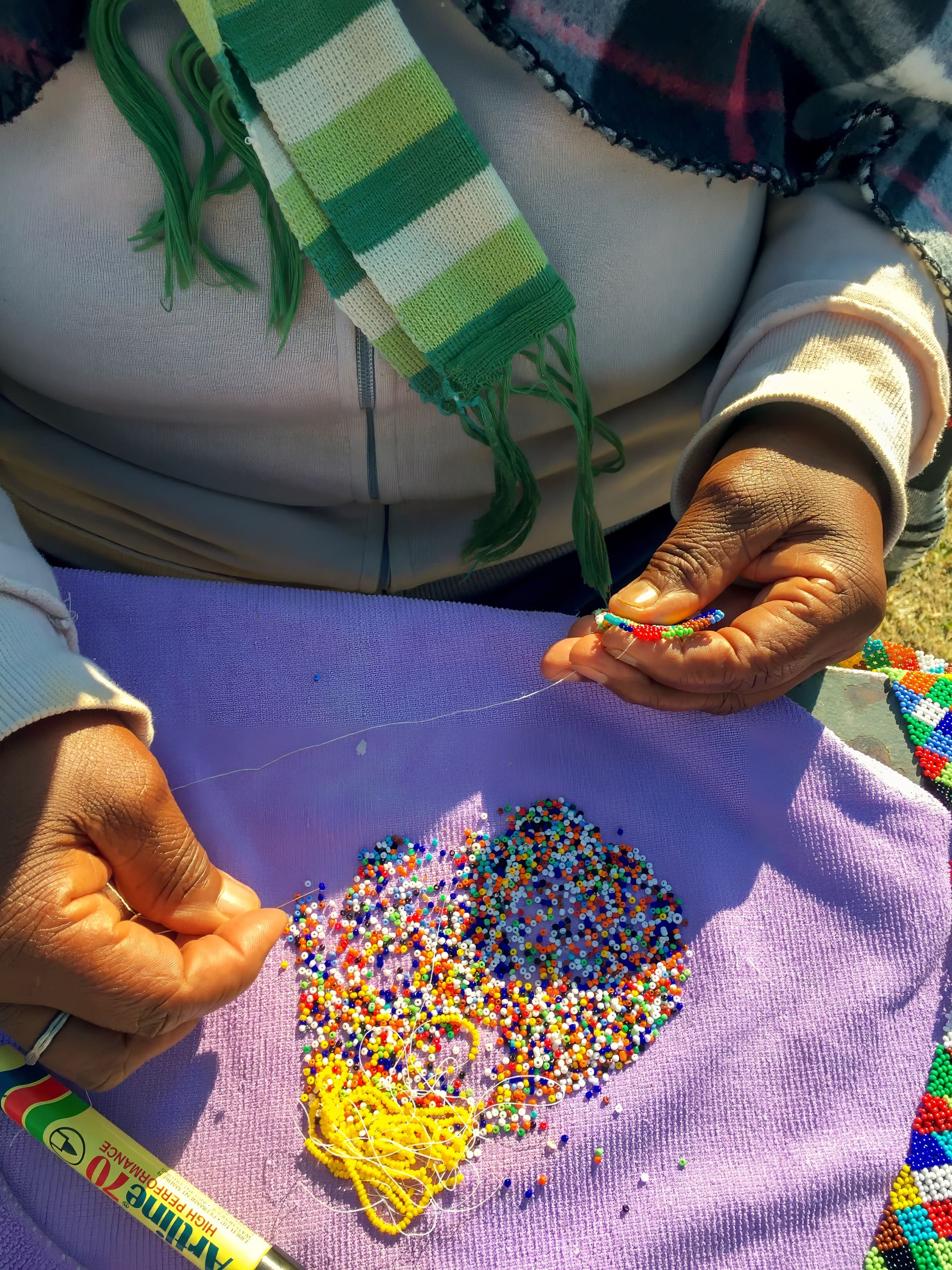 A person’s hands thread colourful beads onto a thread over a lavender cloth scattered with mixed beads. They wear a beige jacket and a green-striped scarf. A tube of glue and finished beadwork are visible on the cloth. The scene is sunlit.