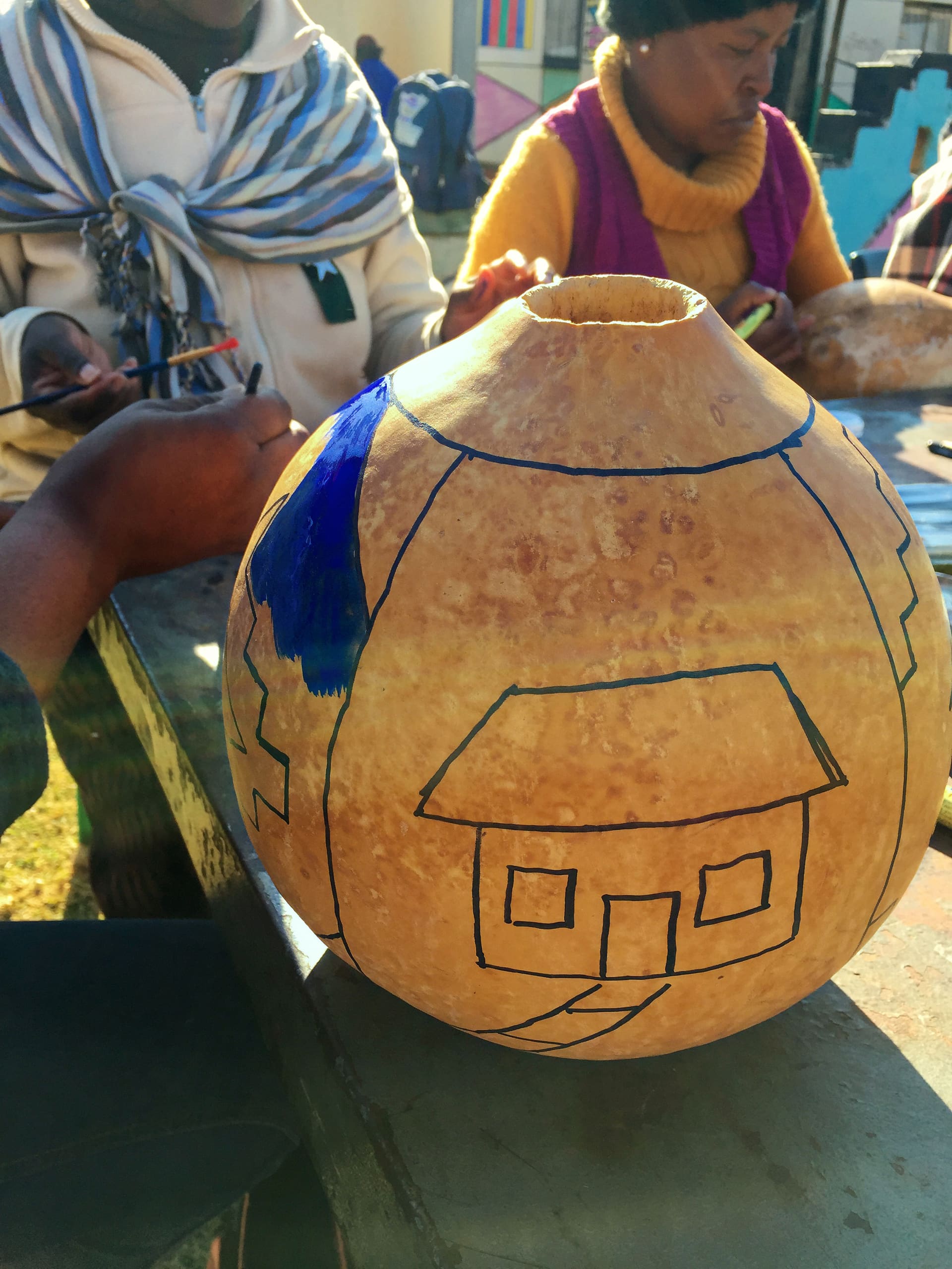 A close-up of a dried gourd with simple house outlines drawn in black marker; a person is painting part of the design blue. In the background, two people sit at a table, focused on their own work, wearing warm clothing and scarves.