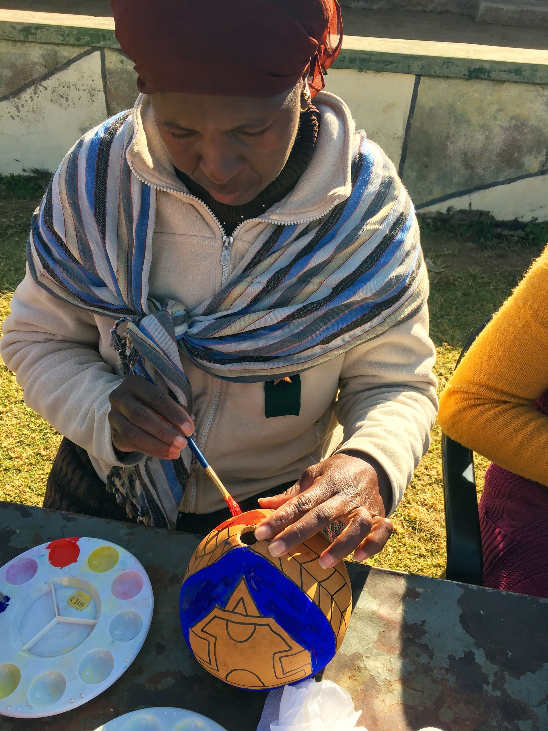 A woman wearing a headscarf, striped shawl, and beige jacket sits outdoors, painting geometric patterns in blue and red on a round gourd. A paint palette with various colours is beside her on a weathered table, and sunlight casts shadows over her hands.