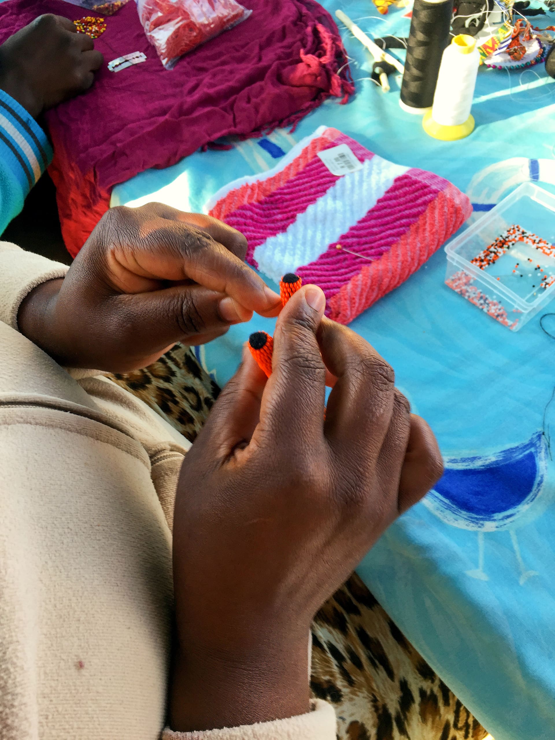 A person with dark skin uses their hands to string black beads onto orange thread. Colourful beads, sewing tools, and fabric are spread out on a blue and pink cloth in the background. The person wears a beige jacket.