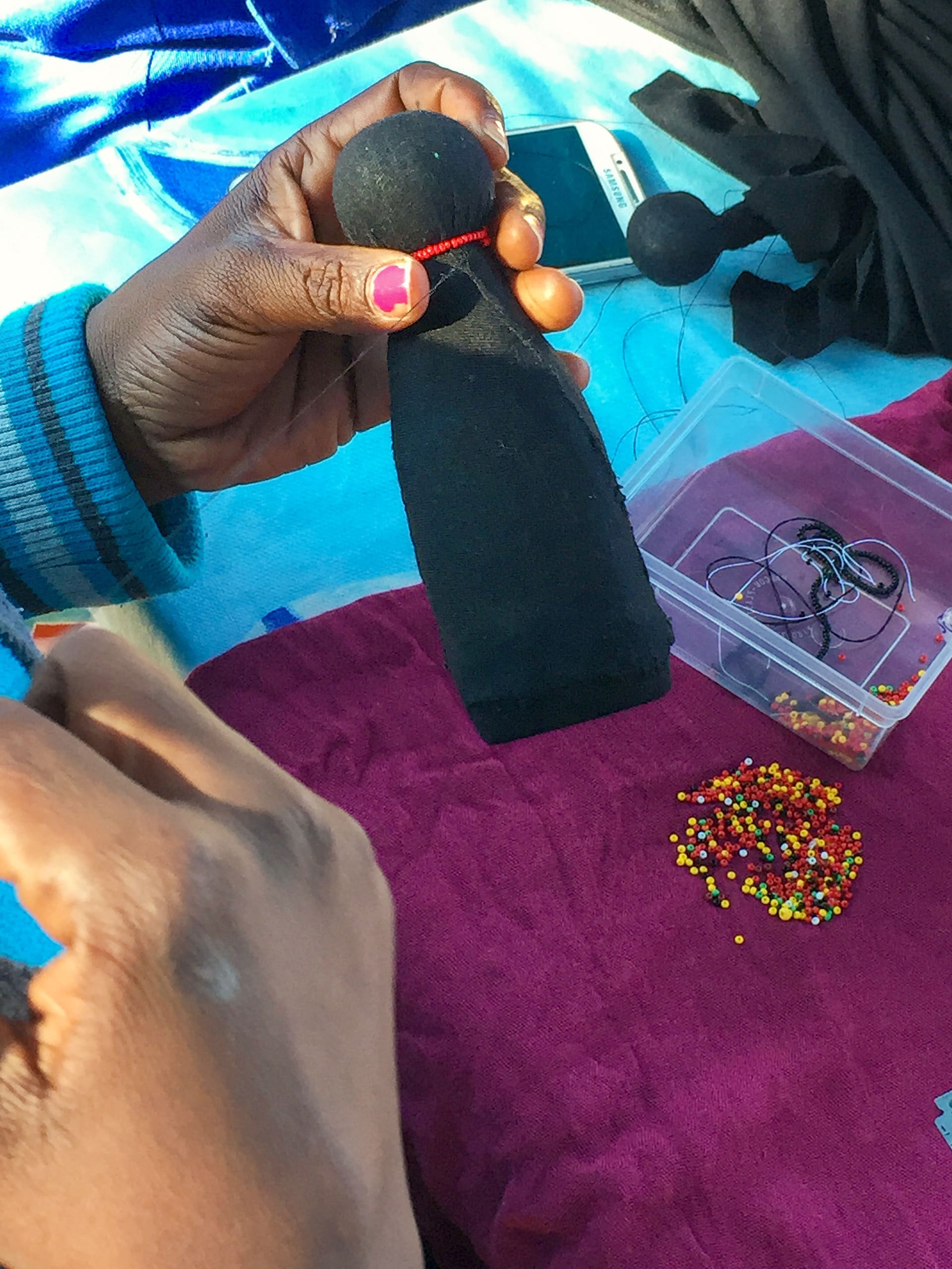 A person with painted nails threads small colourful beads onto a string around the neck of a black cloth doll. Beads and tools are scattered nearby on a burgundy fabric, and another unfinished doll and a plastic box are also visible.