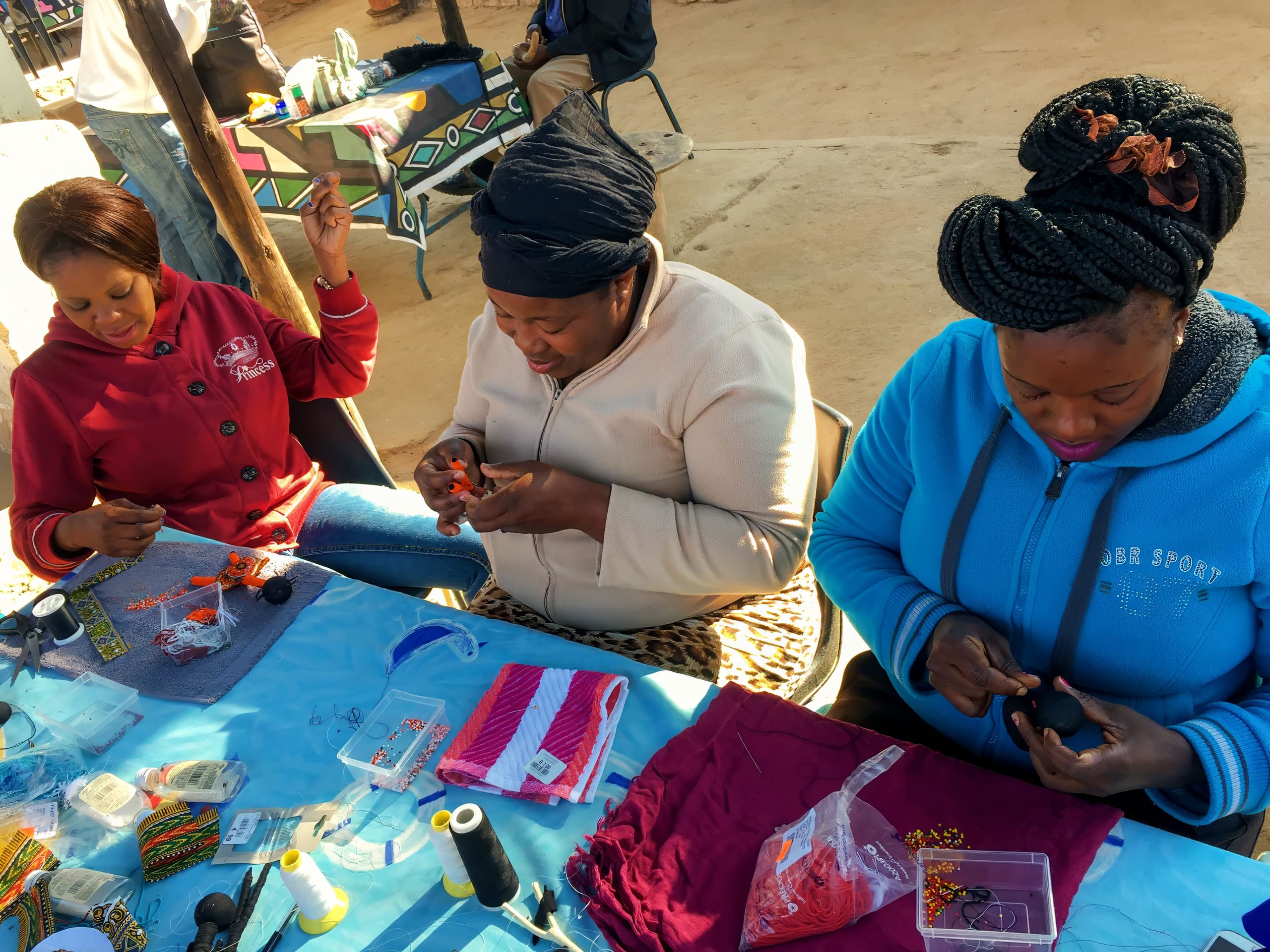 Three women sit at a table covered with beads, thread, and craft supplies, focused on making beaded jewellery. Bright sunlight fills the scene. Each woman works intently with their hands, surrounded by colourful tools and materials.