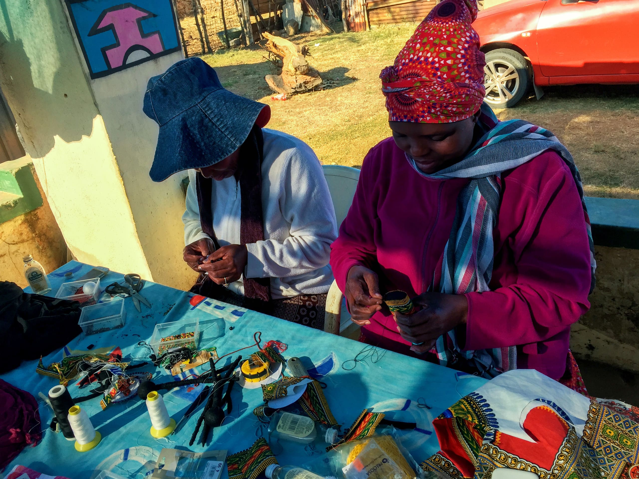 Two women sit at a table covered with colourful beads, thread, and craft supplies, working on beaded jewellery. Both wear head coverings and warm clothes. Sunlight casts shadows across the table. In the background, there’s a red car and a wall with a painted mural.