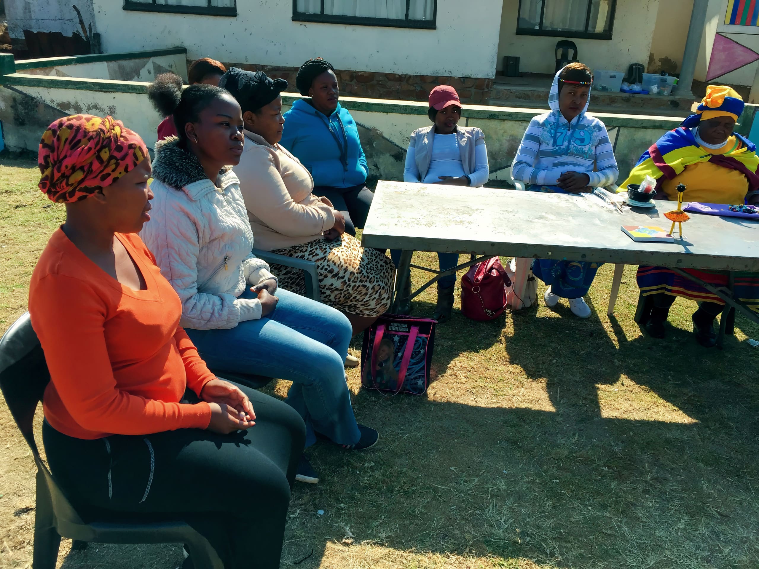 A group of women sit outdoors on plastic chairs around a rectangular table. They appear to be engaged in a discussion or meeting. Some women wear headscarves and brightly coloured clothing. The scene is sunny, with a building and windows in the background.
