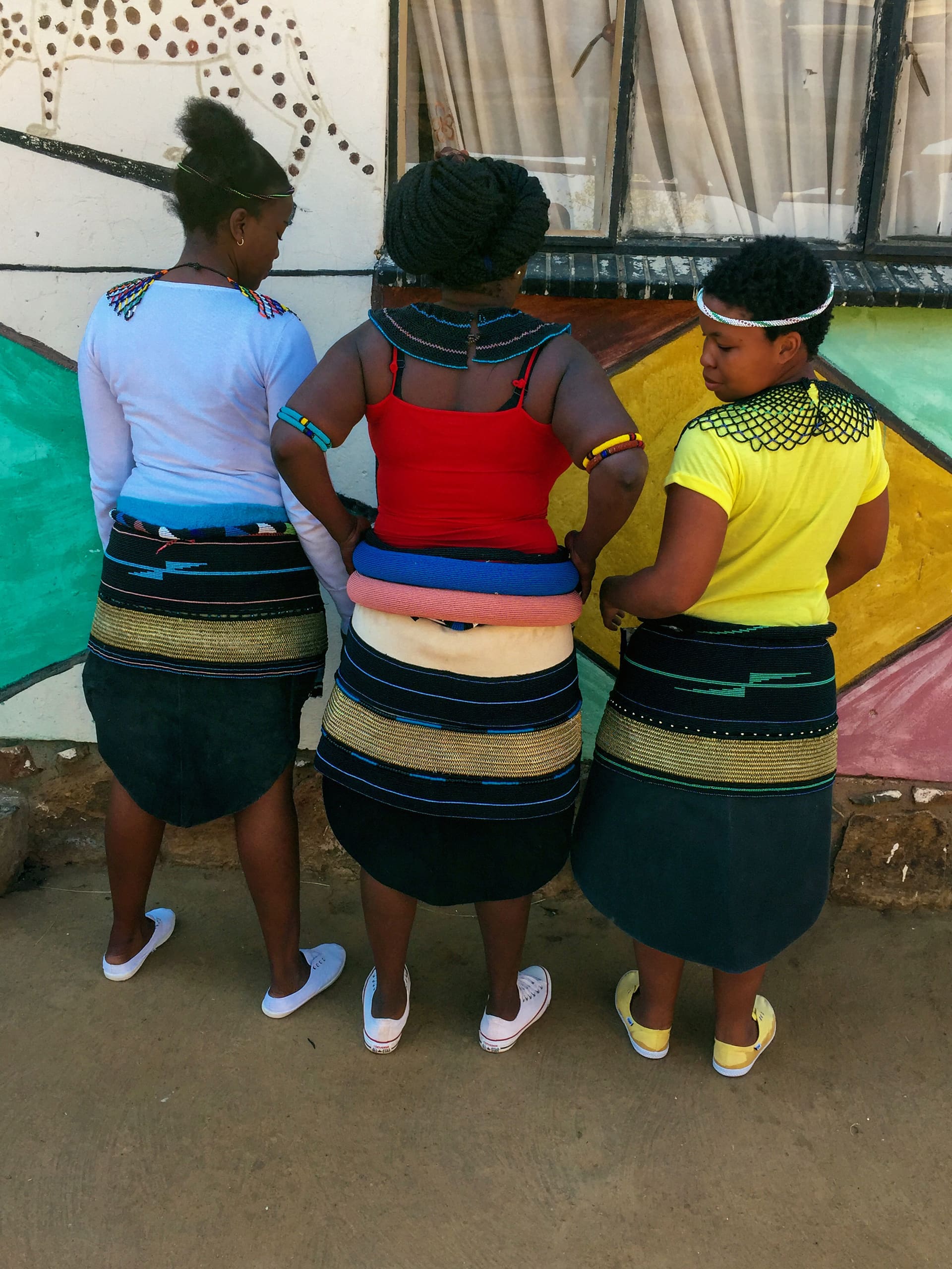 Three women stand with their backs to the camera, wearing traditional beaded skirts and accessories. Their colourful tops—white, red, and yellow—contrast against a painted wall with geometric patterns and a window with sheer curtains.