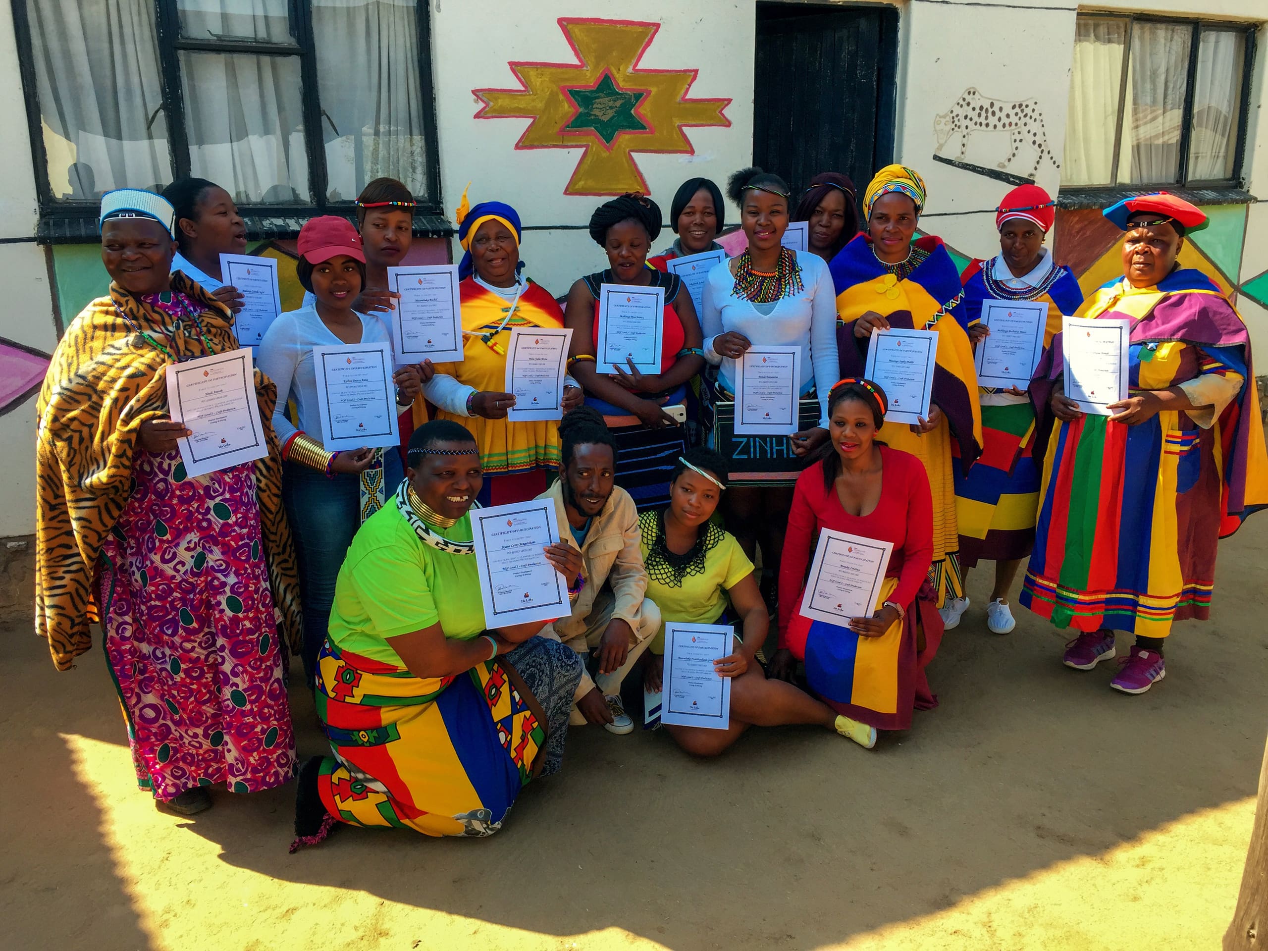 A group of people, mostly women, pose outdoors holding certificates. They wear brightly coloured traditional African clothing and stand in front of a decorated building. Everyone is smiling, celebrating, and facing the camera, suggesting a graduation or achievement event.