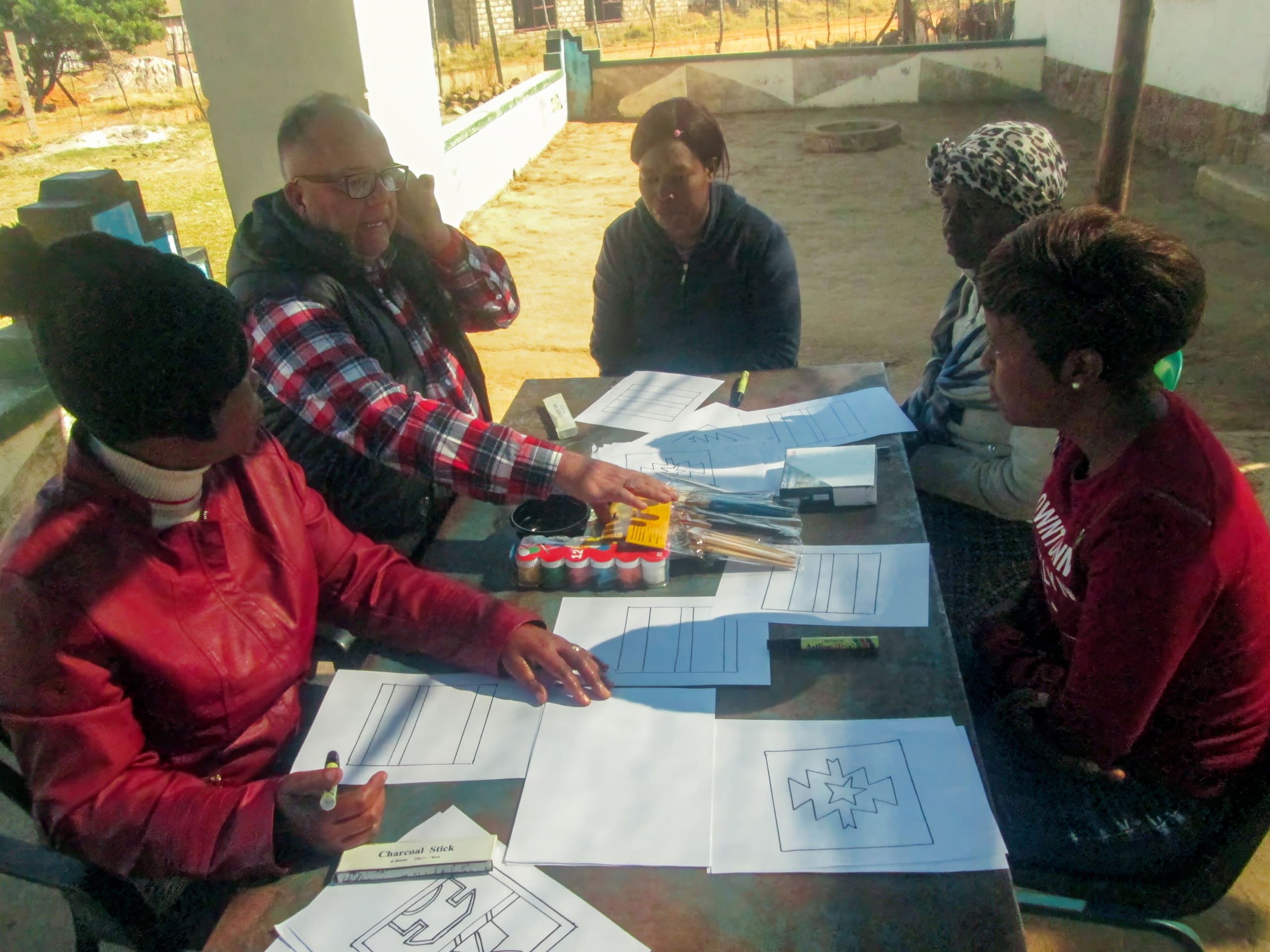 Five adults sit around an outdoor table covered with papers, pens, and art supplies. One man gestures while others listen or write. The group seems engaged in a collaborative activity, possibly a workshop or meeting, on a sunny day.