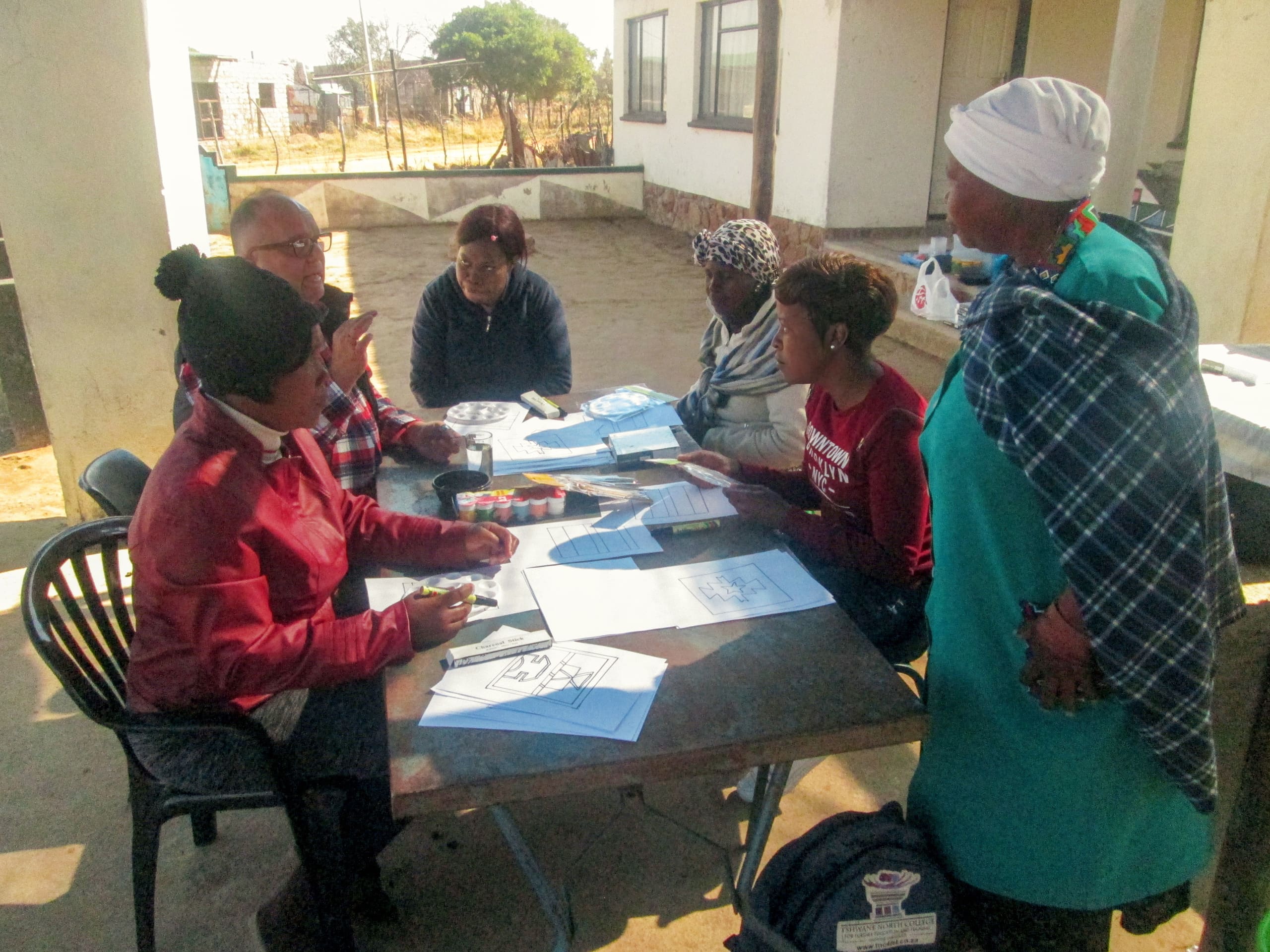 Six women sit and stand around a table outdoors, engaged in discussion. Papers and coloured pencils are spread across the table. The women are dressed warmly; some wear hats or headscarves. Buildings and a garden are visible in the sunny background.