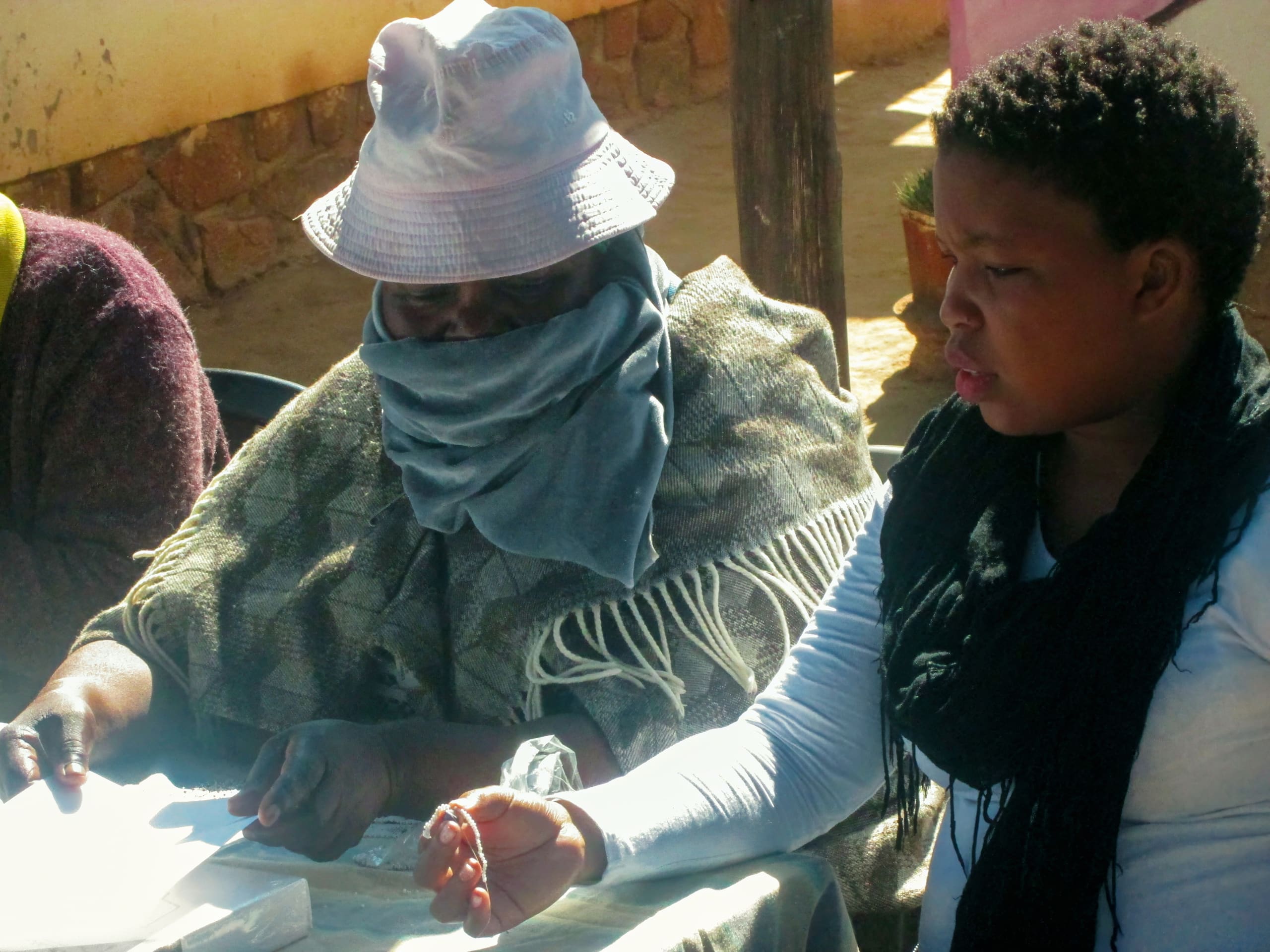 Two people sit side by side at a table. The person on the left wears a bucket hat and has a scarf covering their lower face, whilst the person on the right, a child, looks ahead. Papers and hands are visible on the table in sunlight.