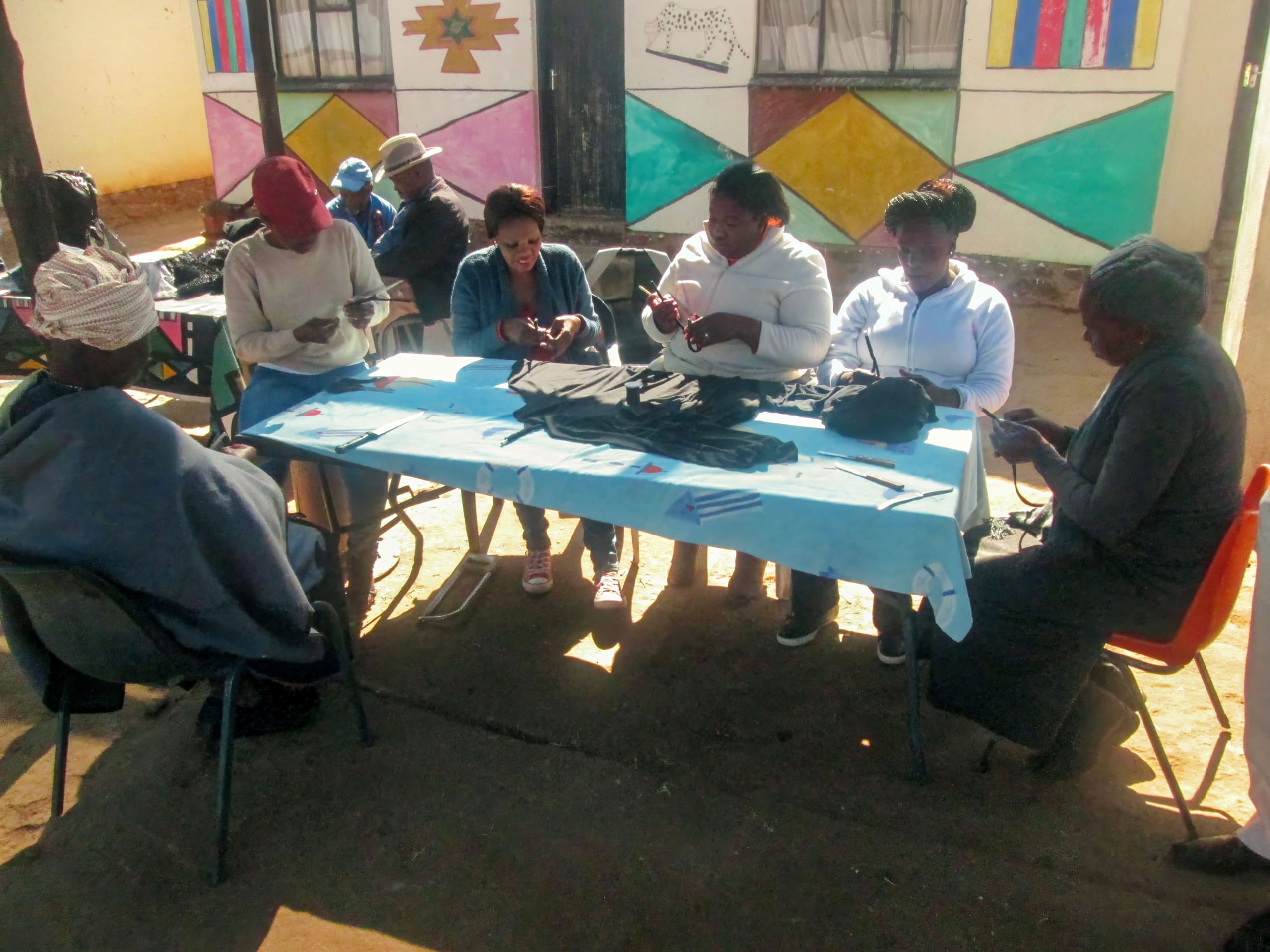 A group of people sit around a blue-covered outdoor table, weaving or crafting items with dark materials. They are focused on their hands. The background includes a mural on a building wall and bright sunlight.