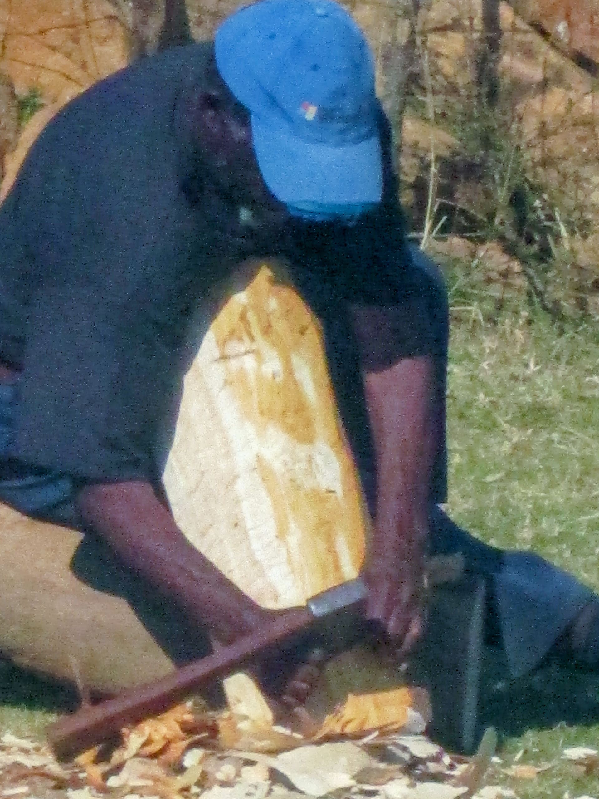 A man in a blue cap sits outside on grass, using an axe to carve or split a large piece of wood. Wood shavings gather around his hands and the log. The background is out of focus, showing earth and vegetation.