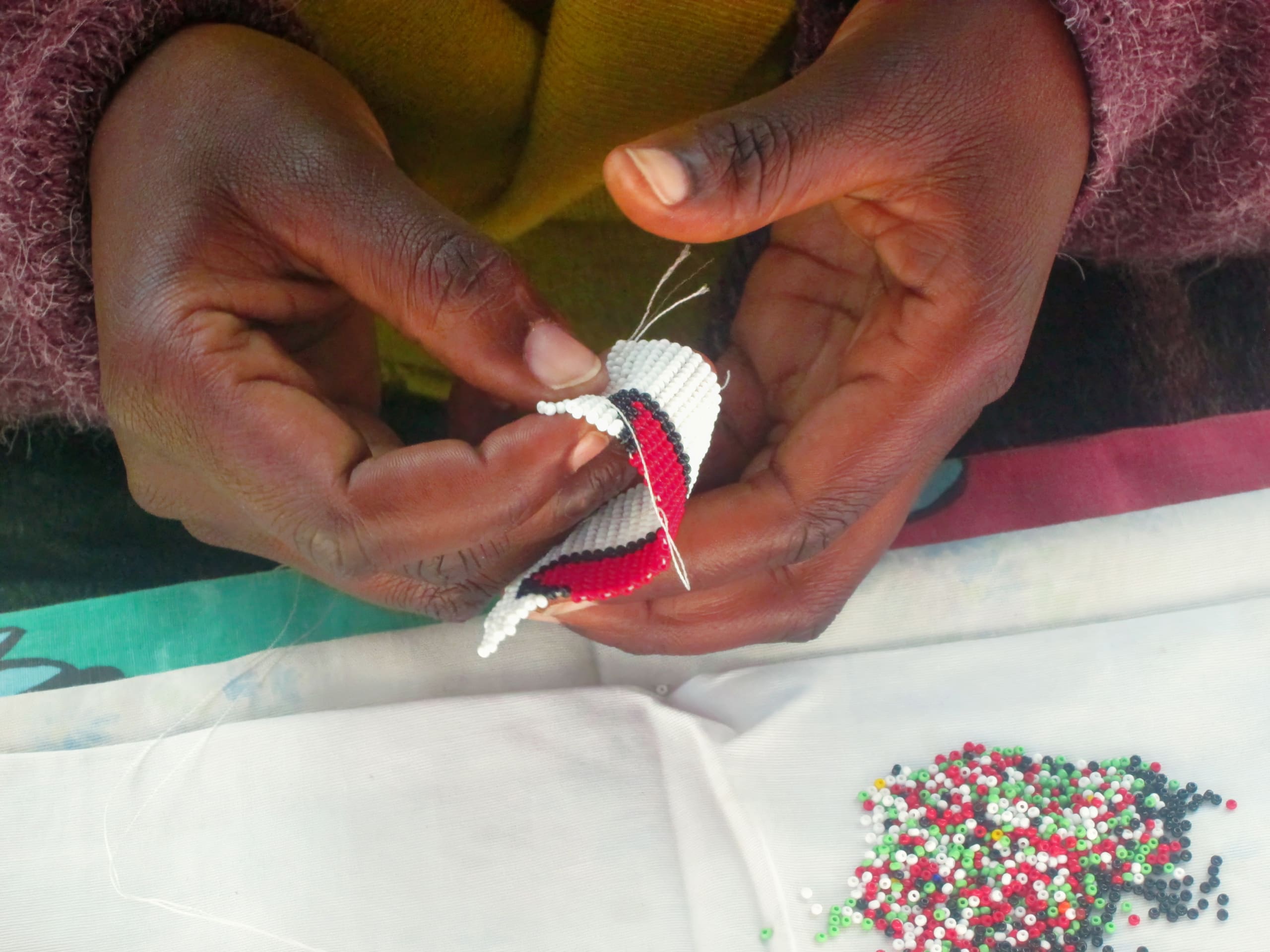 A person with dark skin and a brown jumper uses both hands to weave small red, white, green, and black beads together, forming a patterned piece. A cluster of loose beads in the same colours rests on a white cloth below their hands.
