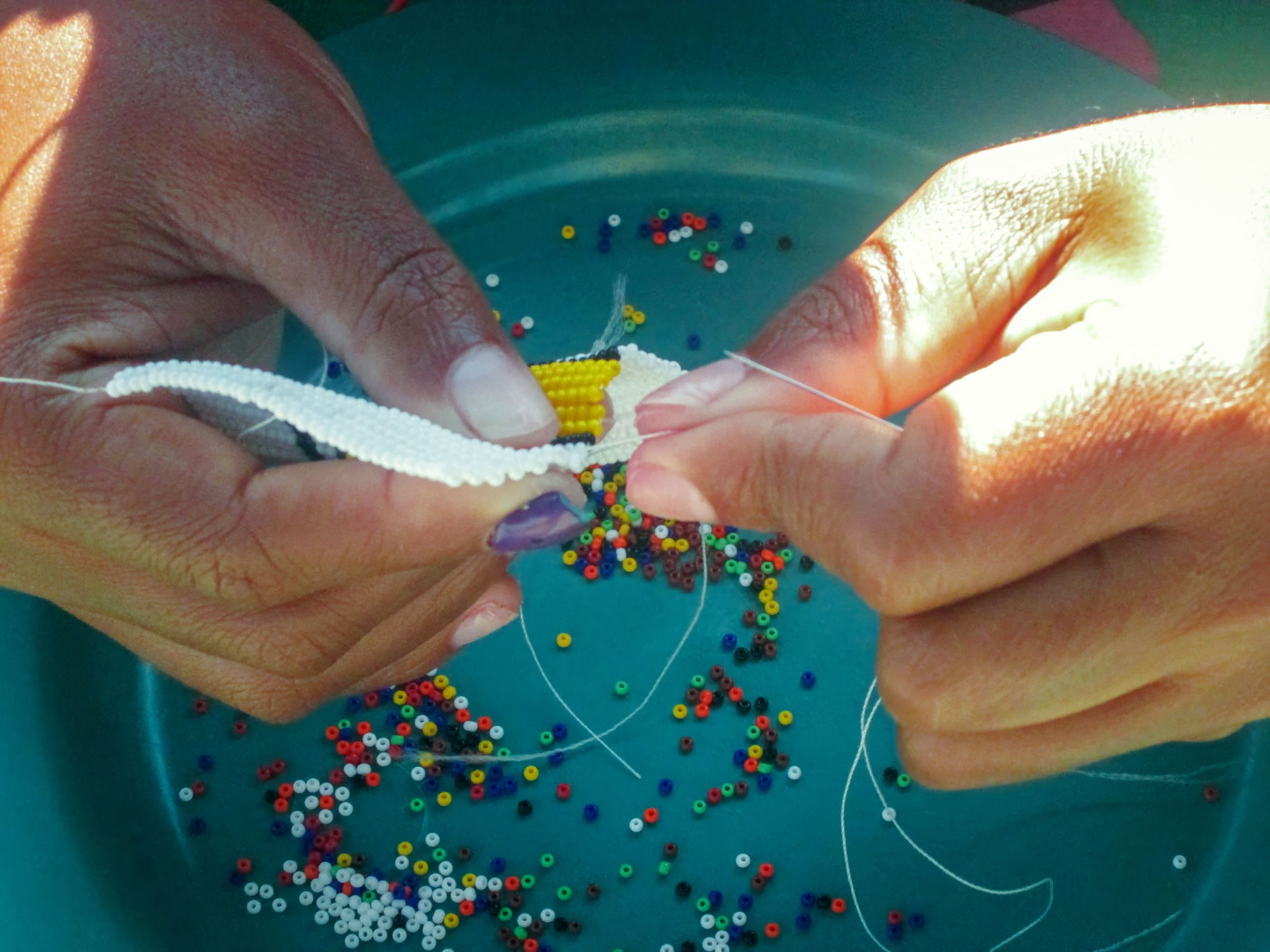 Close-up of two hands threading a needle through white and yellow beads to create beadwork. Multicoloured beads are scattered on a green surface below, with several loose threads visible around the hands. The focus is on the intricate craftwork.