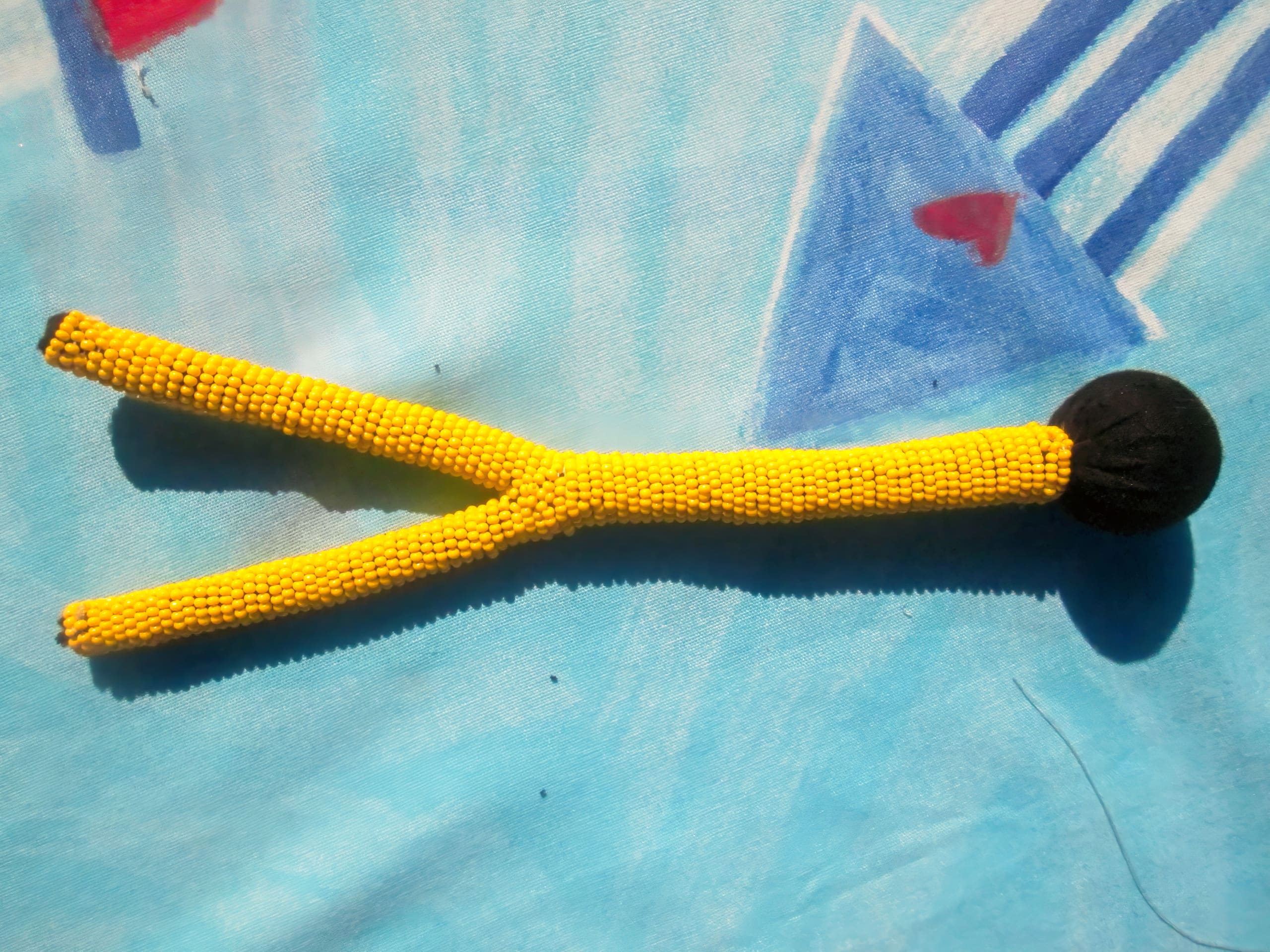 A yellow Y-shaped object made of small beads with a black round end rests on a blue surface decorated with abstract triangle and heart shapes. Shadows are visible, and the background has a soft, textured appearance.