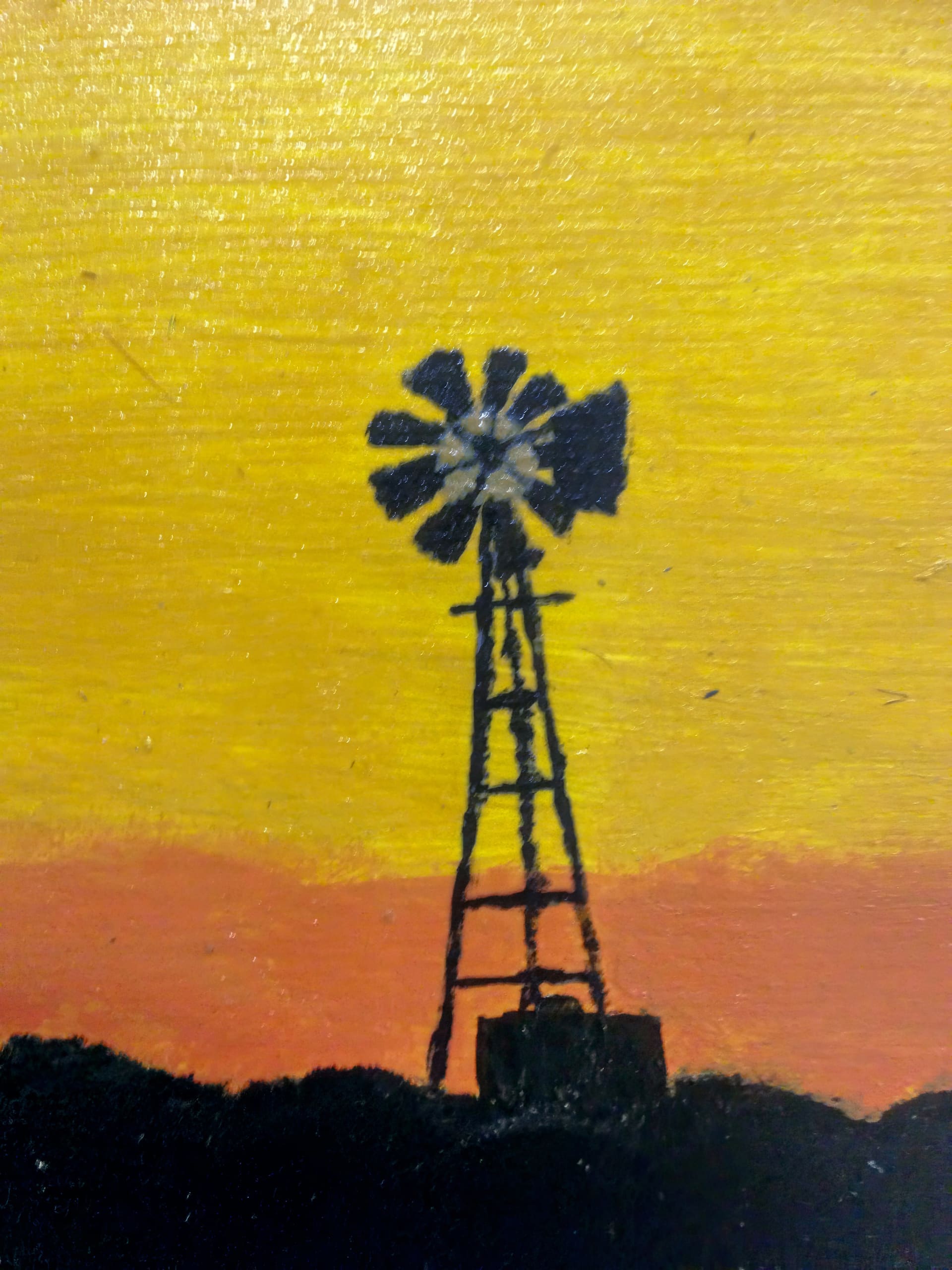 A silhouette of a windmill stands against a vibrant sunset sky, with shades of yellow, orange, and red blending together. The windmill’s sails and tower are dark, contrasting with the colourful, glowing background and the shadowy ground below.