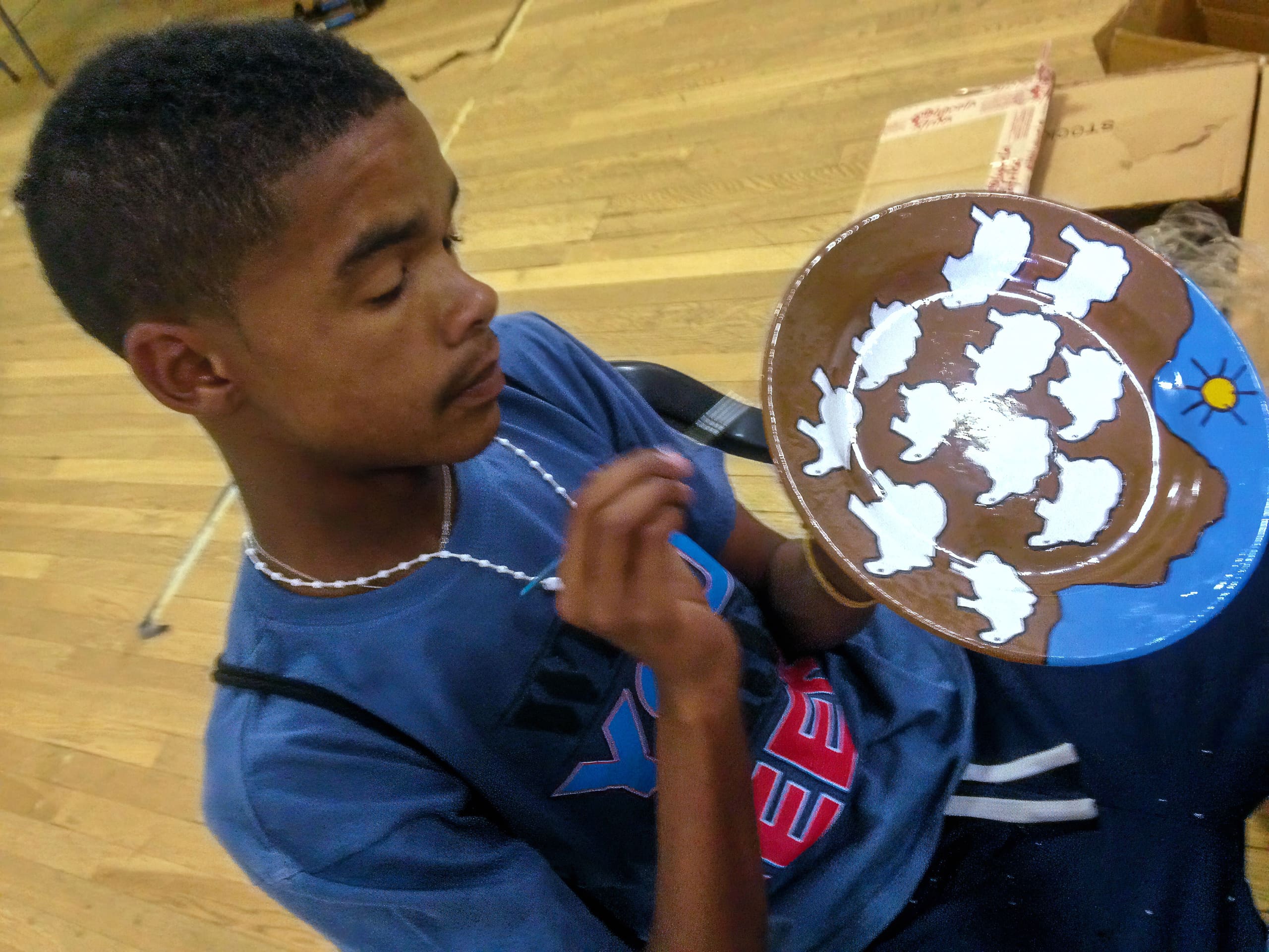 A young boy with short hair, wearing a blue shirt and necklace, holds and carefully paints a brown plate. The plate features white buffalo silhouettes circling a blue area with a yellow sun. The boy is seated on a wooden floor with boxes in the background.