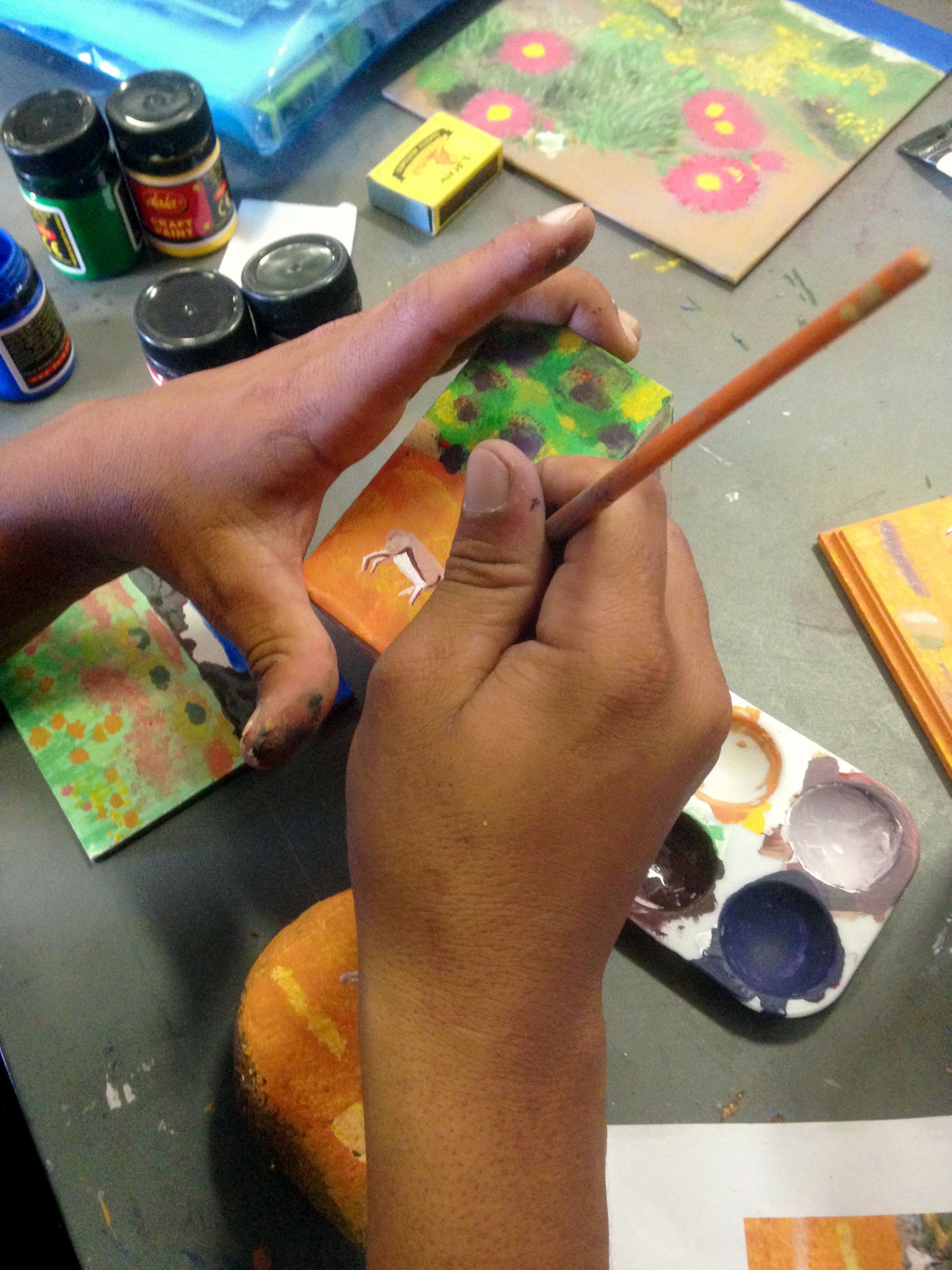 A person with paint-stained hands holds a small canvas and uses a thin brush to paint. Nearby are jars of paint, a palette with various colours, and another painted card with flowers, all on a grey tabletop, suggesting a creative art activity.