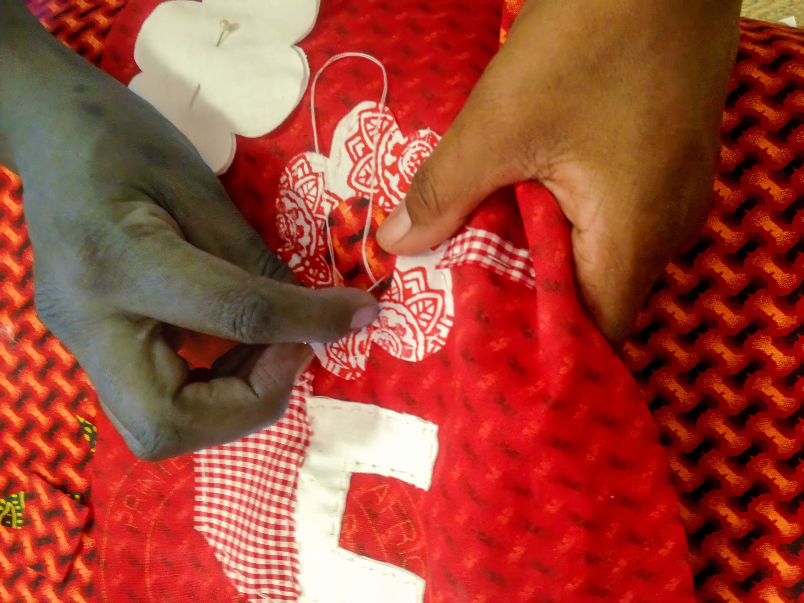 Two hands, one dark-skinned and one light-skinned, sew a red fabric with white and gingham patches. One hand holds the fabric steady, while the other inserts a threaded needle into an embroidered area. The background has a textured, patterned red fabric.