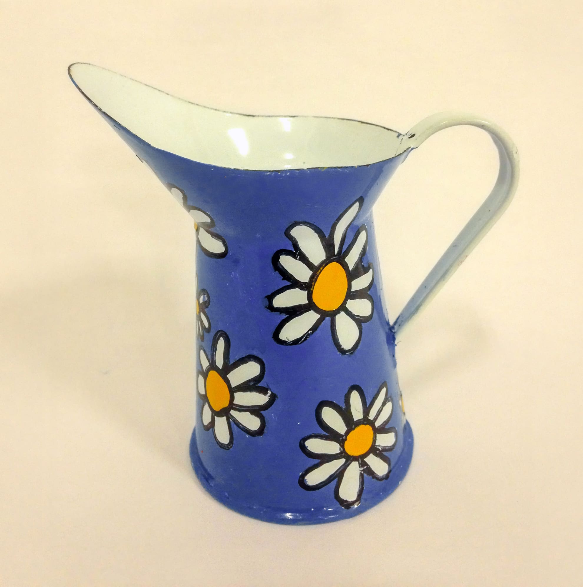 A blue metal jug with a side handle, decorated with white and yellow daisy flowers outlined in black. The inside of the jug is white, and the background is plain and light-coloured.