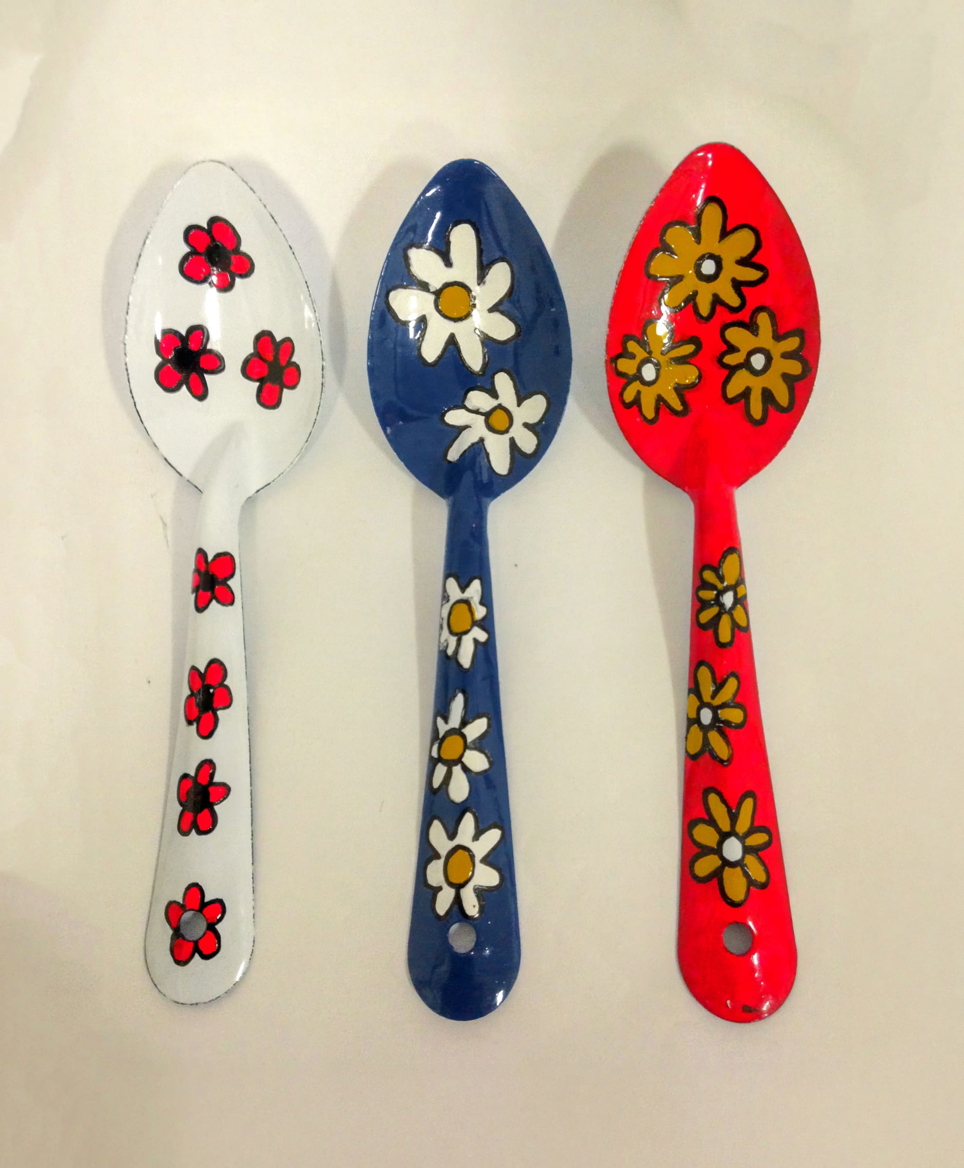 Three ceramic spoons are shown side by side. The left spoon is white with red flowers, the middle is blue with large white and yellow daisies, and the right is red with large yellow and white daisies. All have floral designs and glossy finishes.
