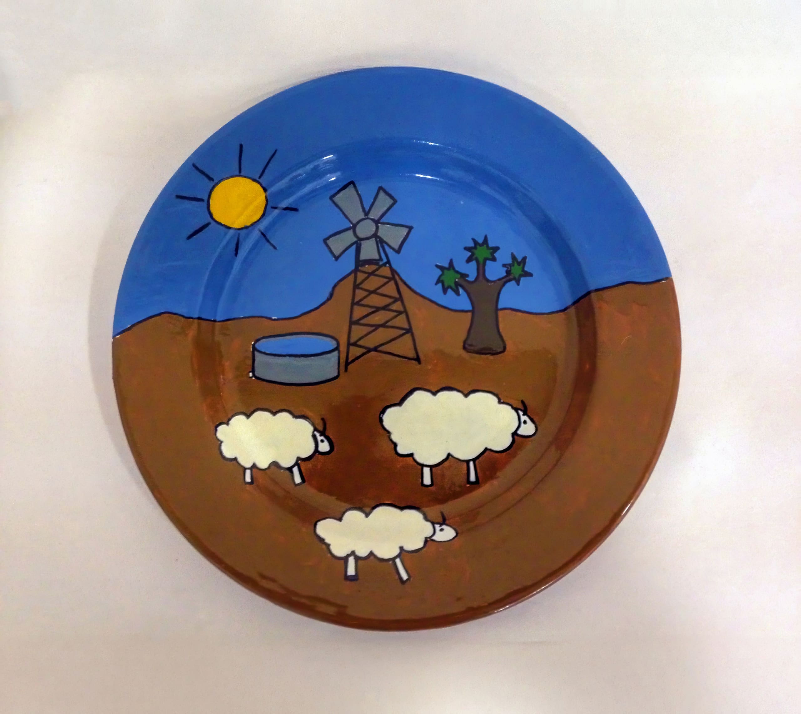 A painted plate shows three white sheep grazing on brown ground, with a tree, a water tank, a windmill, and a bright yellow sun in a blue sky above them. The cartoon scene depicts a rural farm landscape.