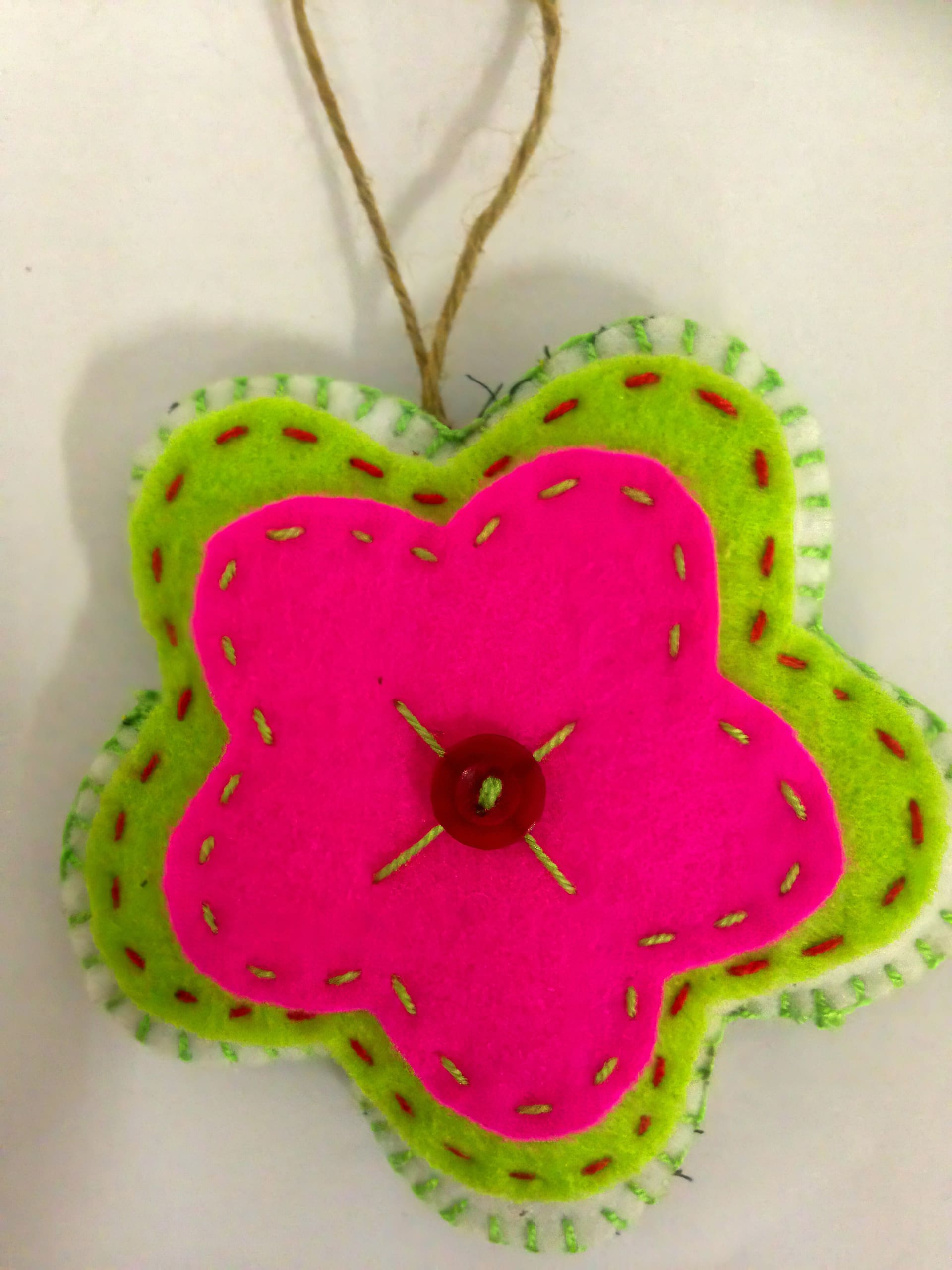 A handmade felt flower ornament with three layers: green, white, and bright pink, sewn together with visible stitching. A red button is attached at the centre, and a loop of brown string is used for hanging. The background is plain white.
