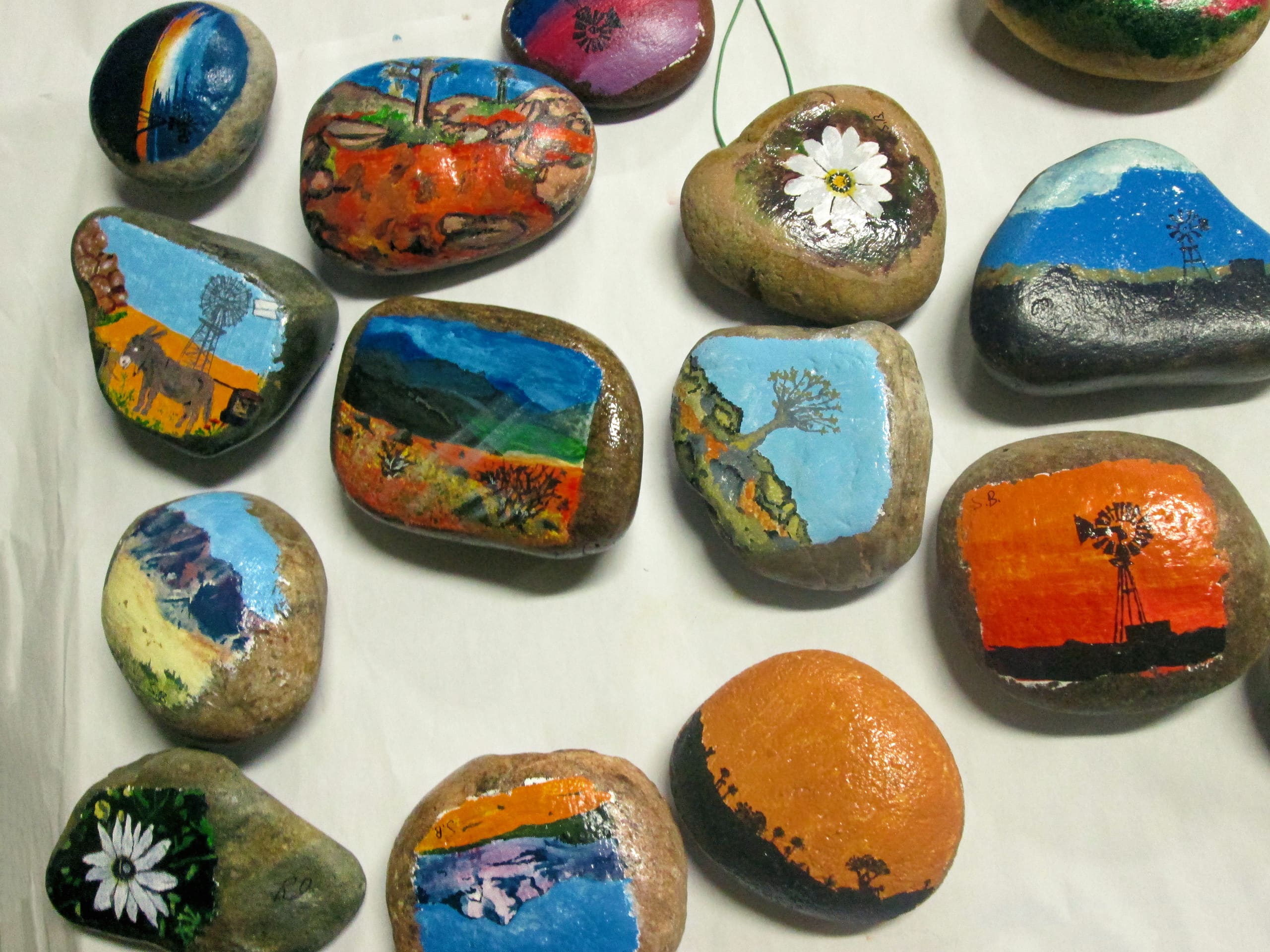 Several smooth rocks are painted with colourful landscapes, daisies, sunsets, windmills, and mountain scenes. The rocks are arranged on a white background, displaying the vibrant outdoor-themed artwork on each stone.