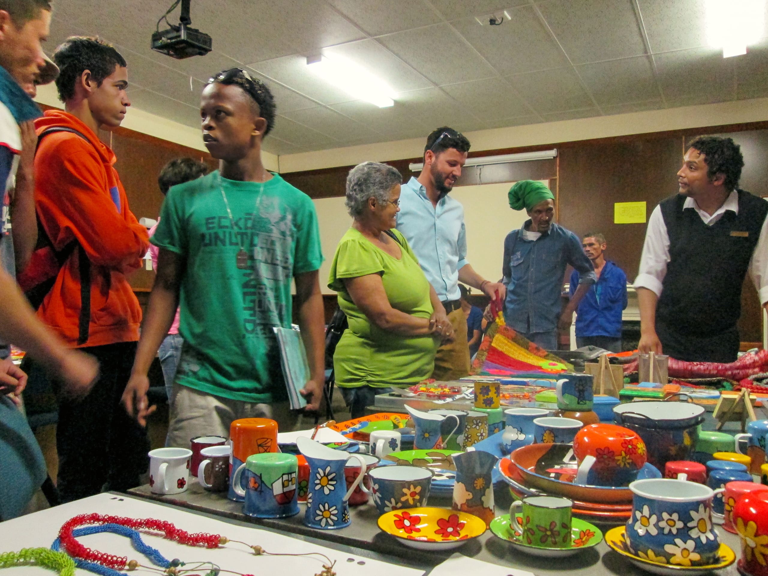 A diverse group of people stands and talks around tables covered with colourful, hand-painted pottery, mugs, bowls, and jewellery at an indoor craft fair. The room has brown walls, fluorescent lights, and a warm, lively atmosphere.