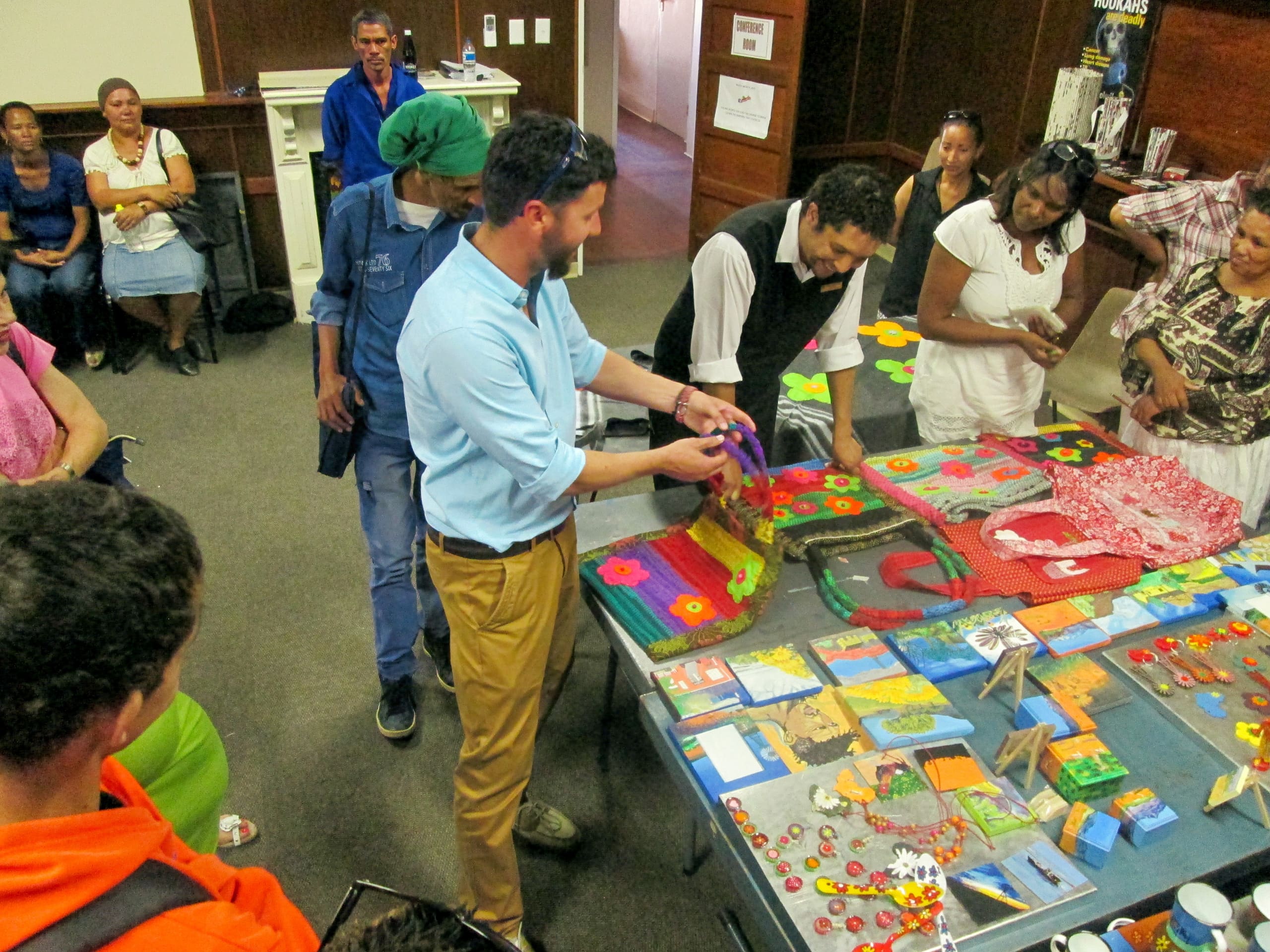 A group of people gather around a table displaying colourful handmade crafts, including textiles with flower patterns and painted tiles. One man in a blue shirt examines a piece, whilst others watch and discuss the items in a brightly lit indoor room.
