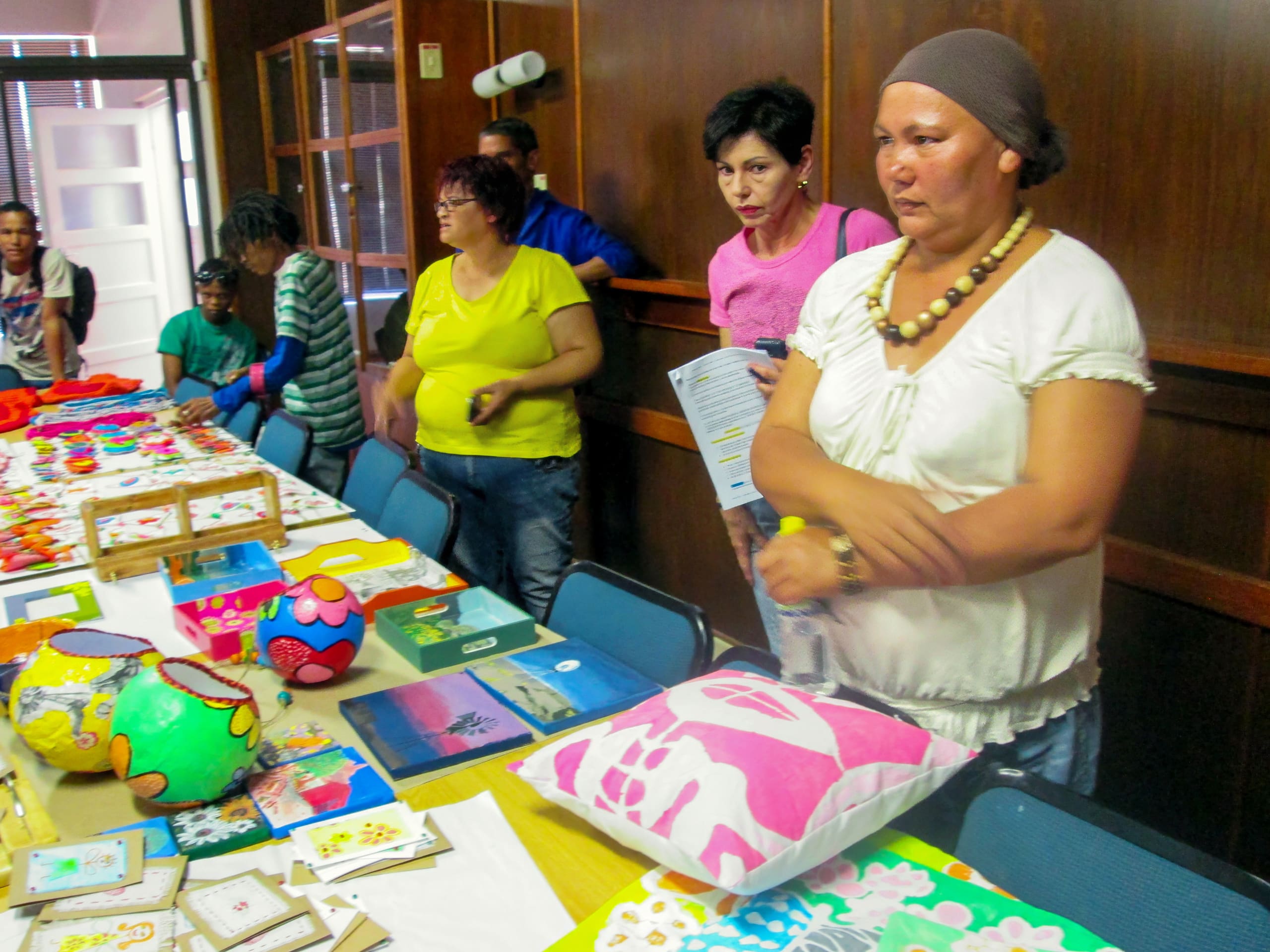 A group of people stands around a table displaying colourful handmade crafts, including painted objects, cushions, and framed artwork. The setting appears to be indoors, with wood-panelled walls and natural light from a door and window in the background.