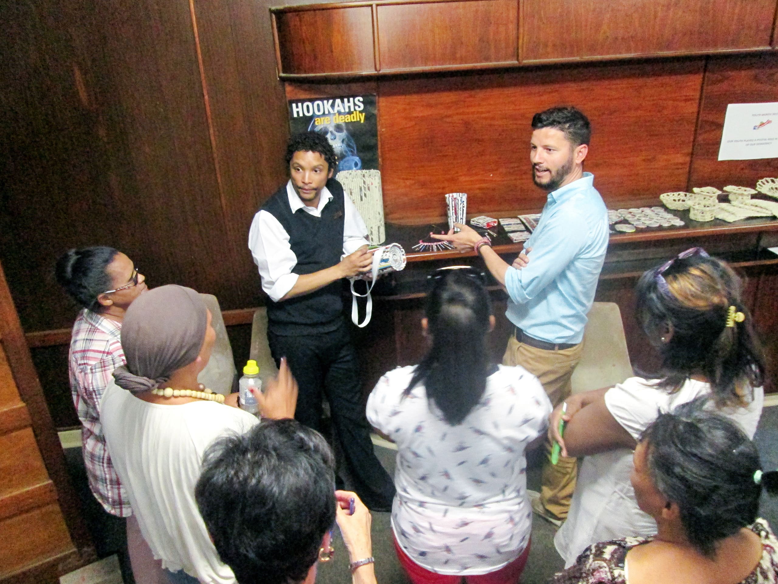 A man performs a rope magic trick for a small group indoors. Another man watches beside him, holding cards. The group, mostly women, stands in a semi-circle, observing. Behind them is a shelf with 