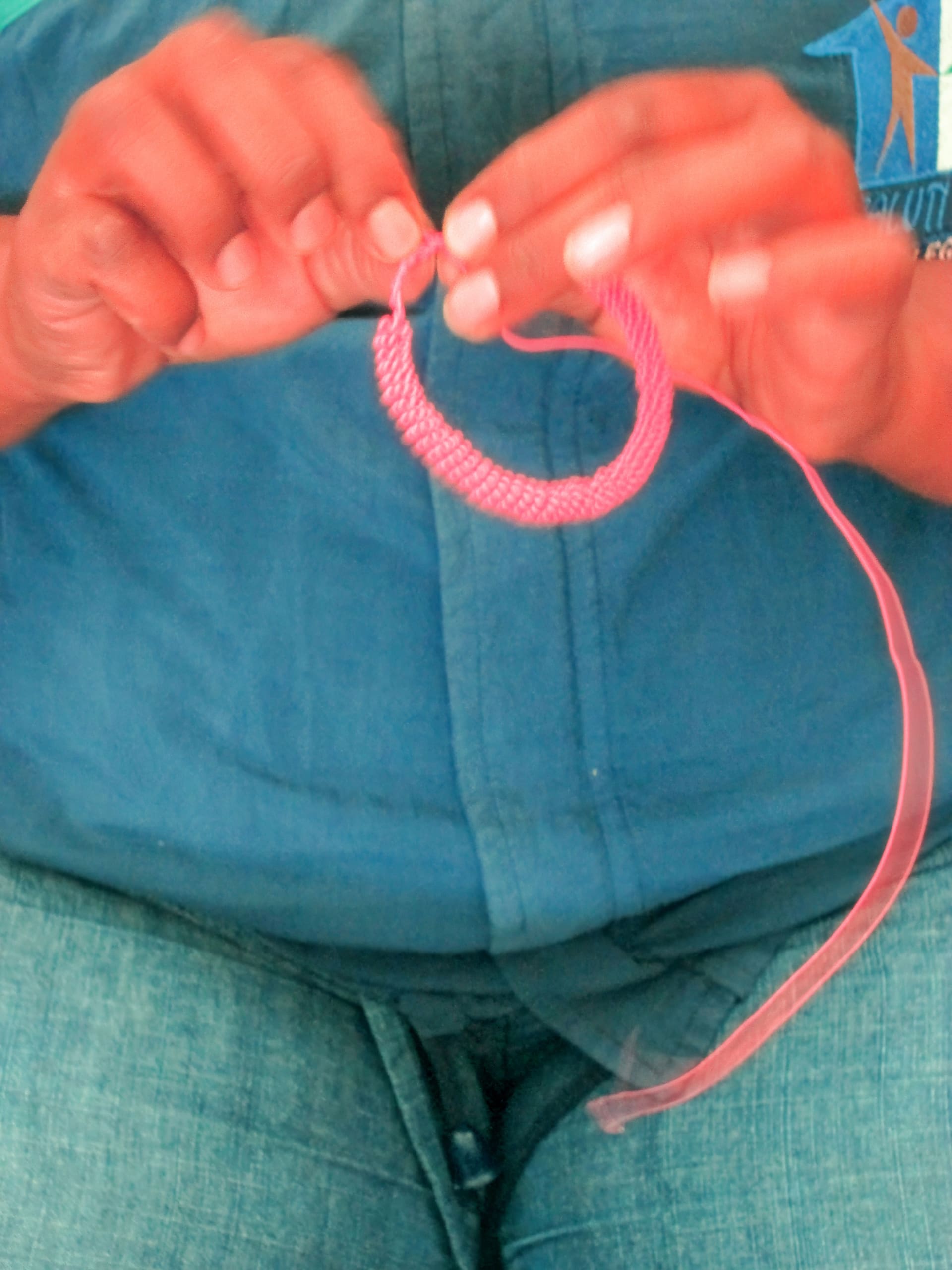 A person in blue clothing and jeans is holding and weaving a circular pink beaded object with their hands, threading a string through the beads. The focus is on the hands and craft, not showing the person's face.