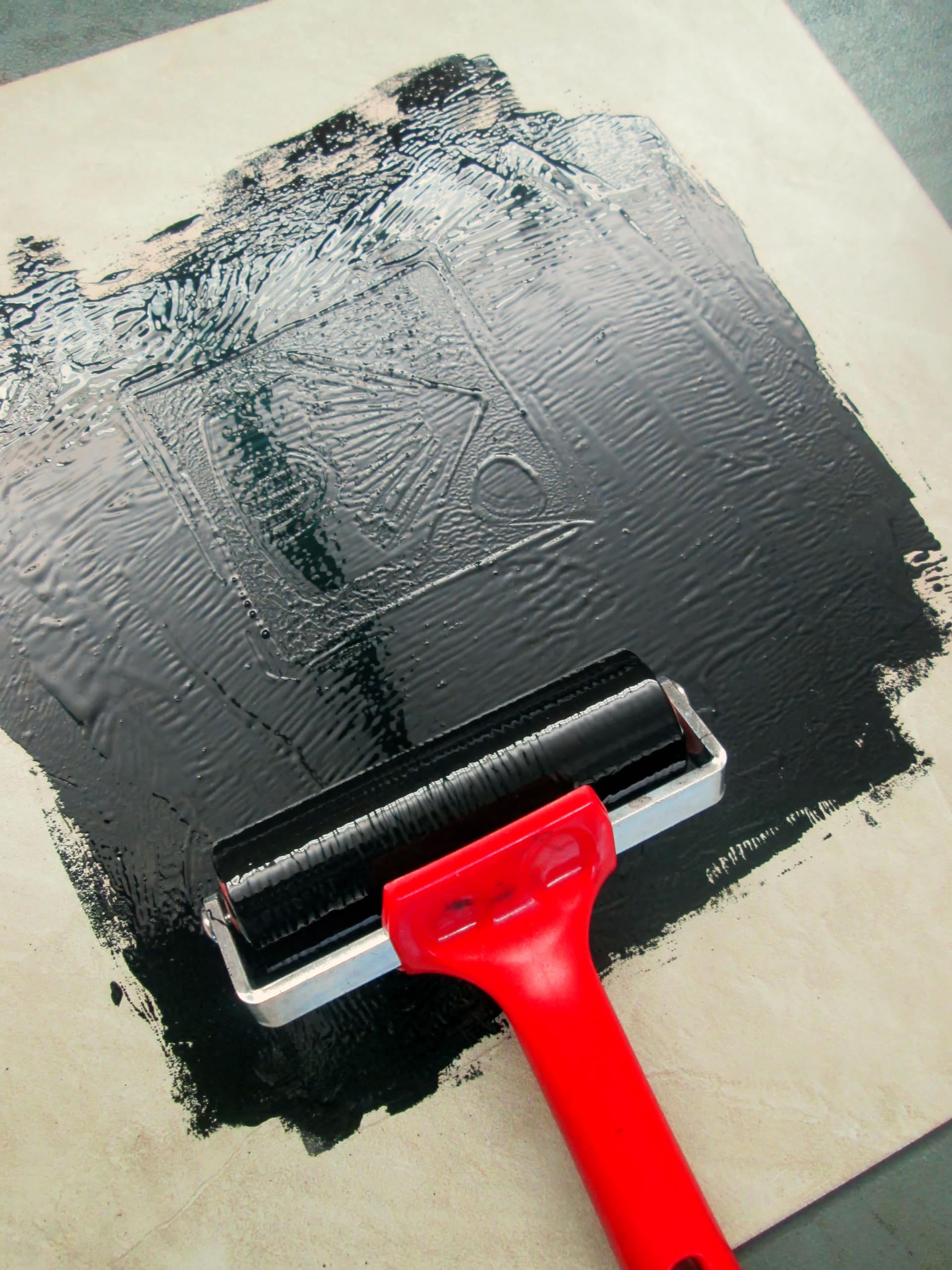 A red-handled roller spreads black ink over a carved lino block on a white surface, preparing it for printmaking. The block displays a partially visible design, and the ink appears thick and glossy.