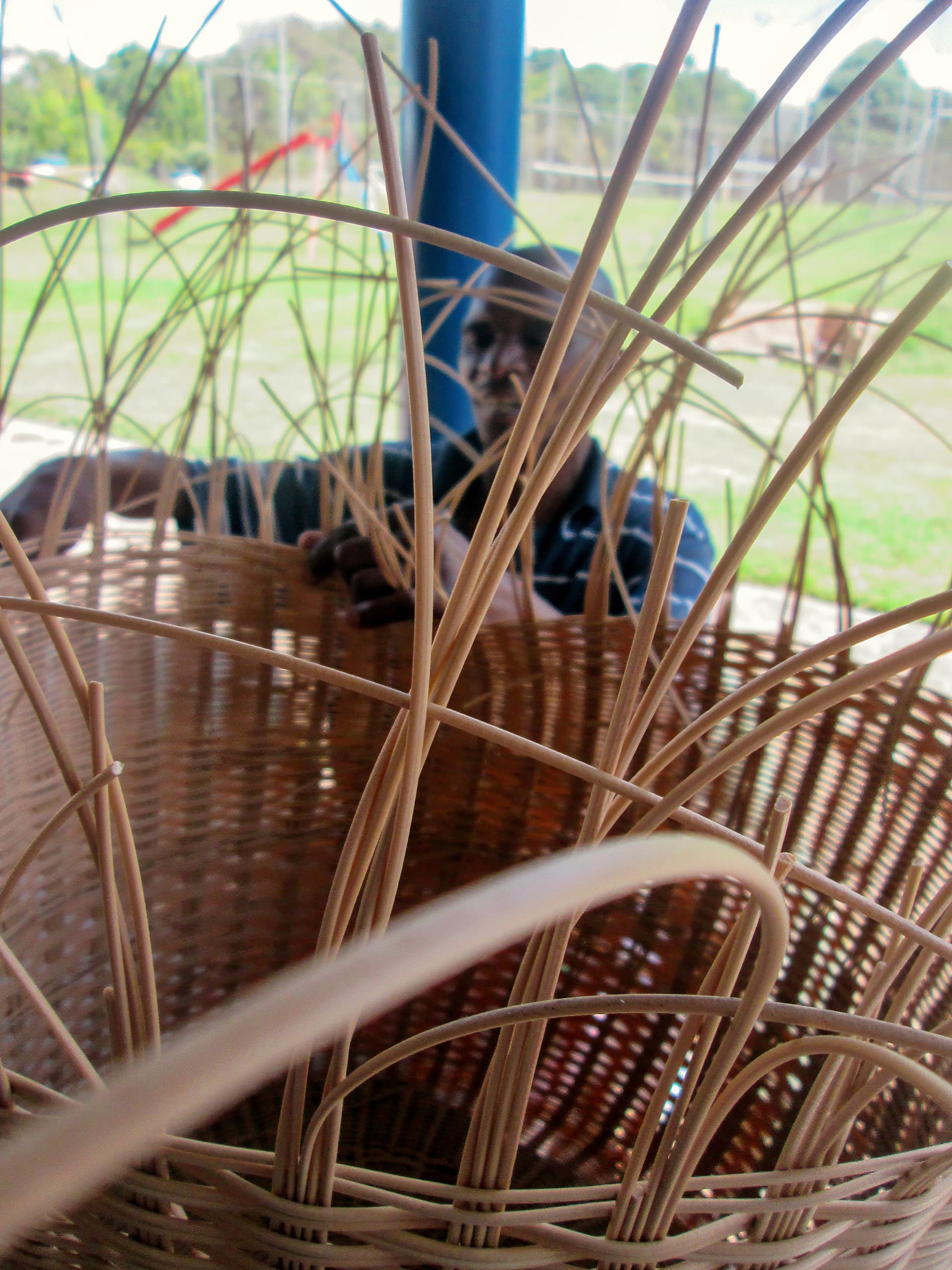 A close-up view of a partially woven wicker basket with long, thin reeds sticking up. In the background, a person’s face and hands are visible, focused on weaving the basket. The setting appears to be outdoors with blurred green grass and structures behind.